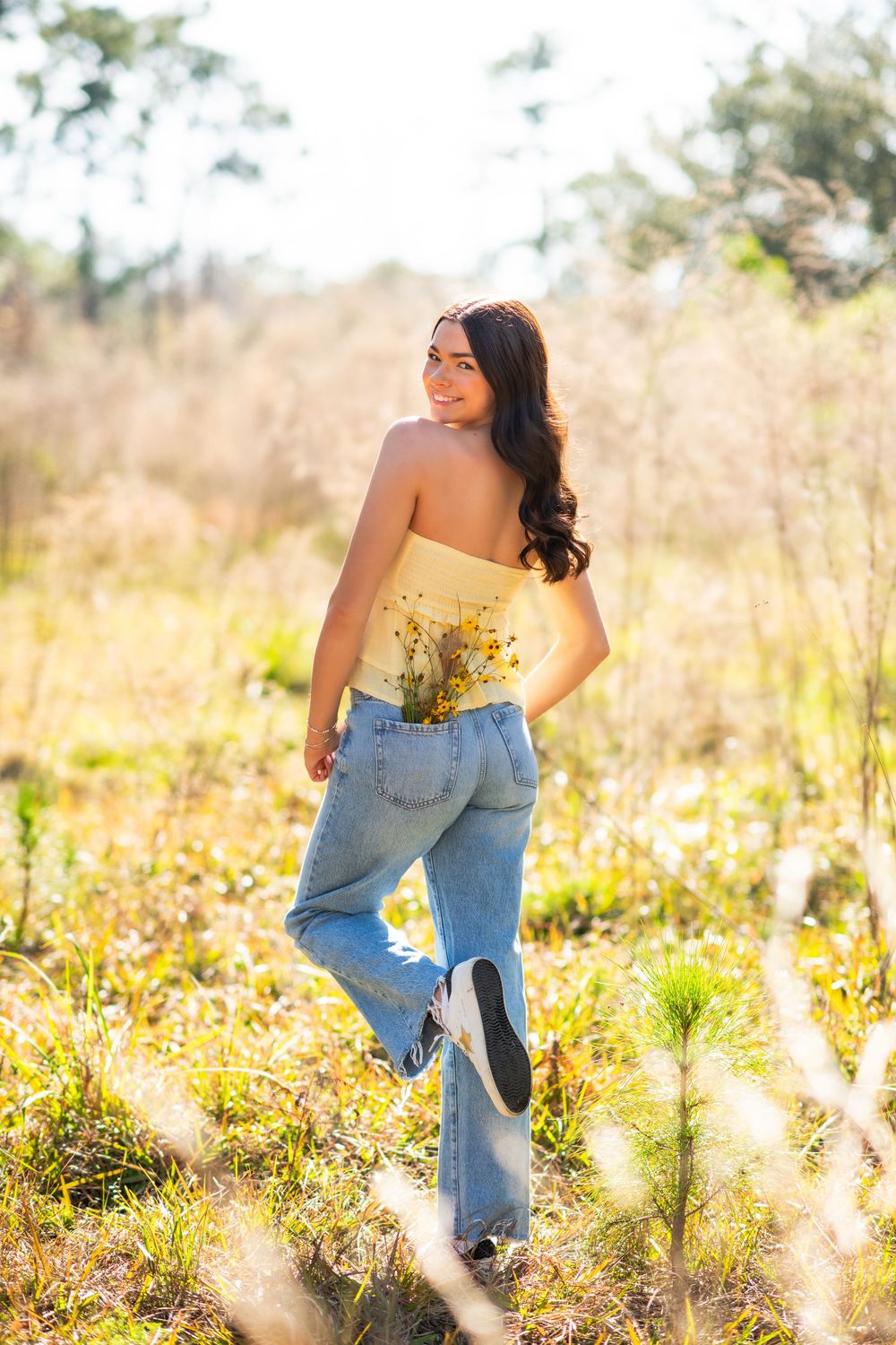 Young woman in yellow tube top and jeans holds wildflowers while walking through a sunny meadow, smiling over her shoulder.