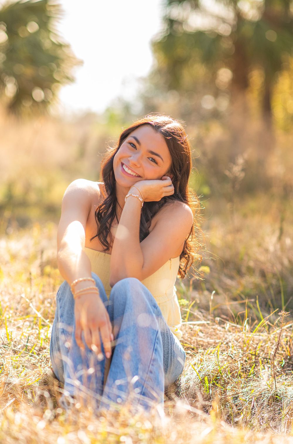 Young woman sitting in sunlit field, smiling warmly in yellow top and jeans, with golden bokeh background.