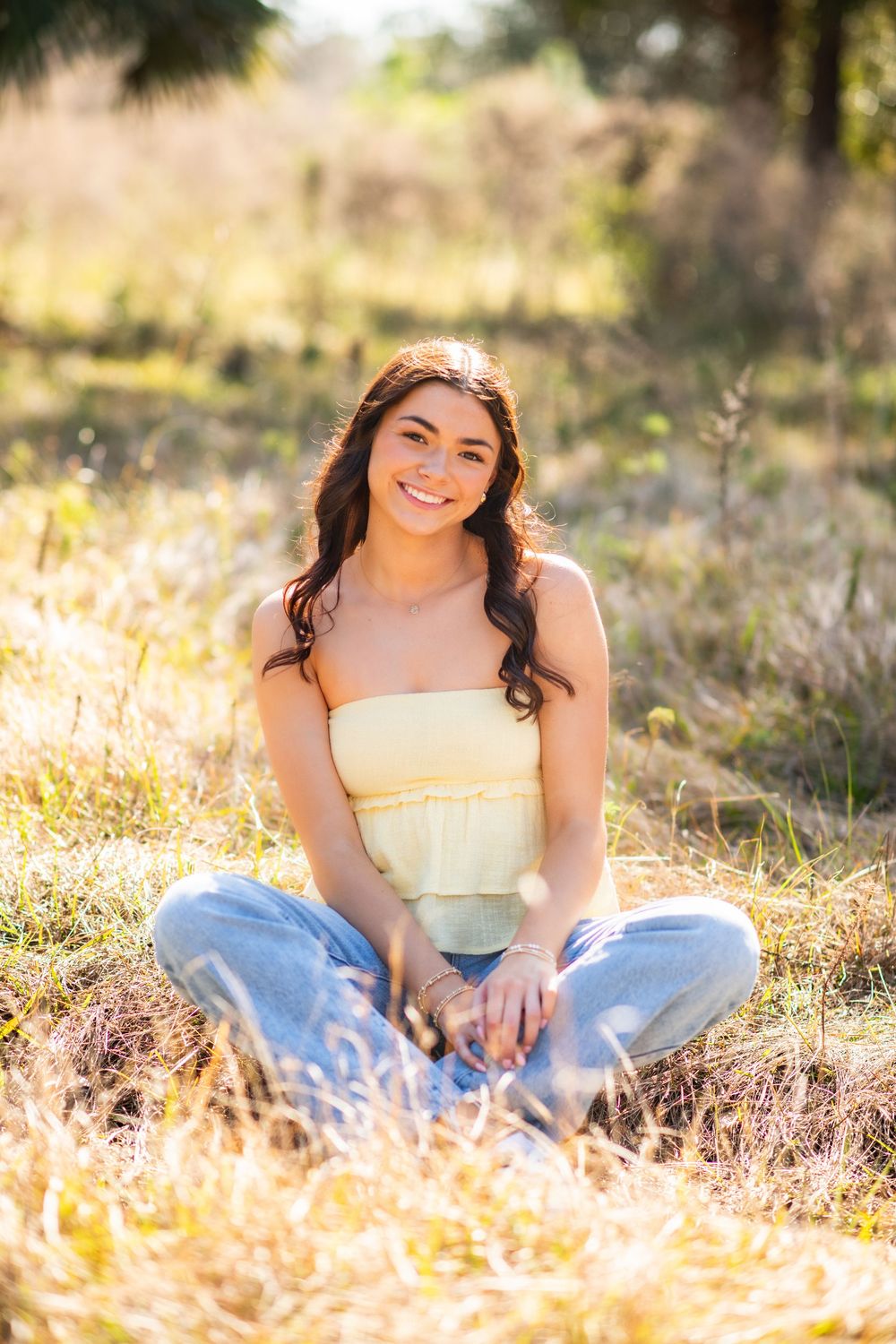 Young woman in yellow tube top and jeans sitting cross-legged in a sunlit field smiling.