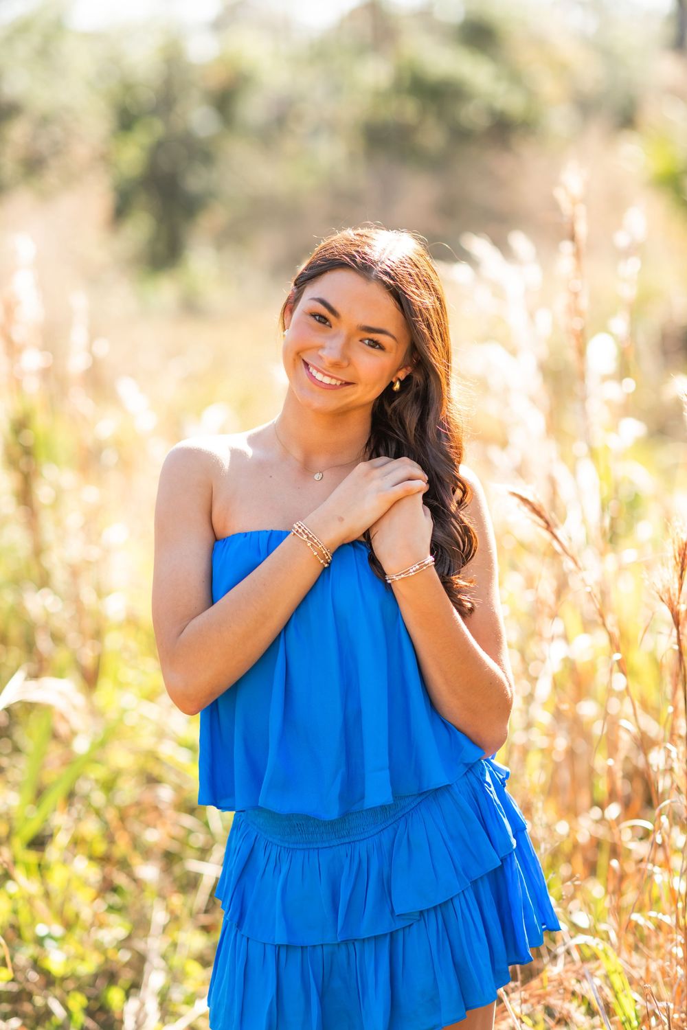 Smiling young woman in a blue strapless ruffled dress standing in a sunlit field with tall golden grasses.