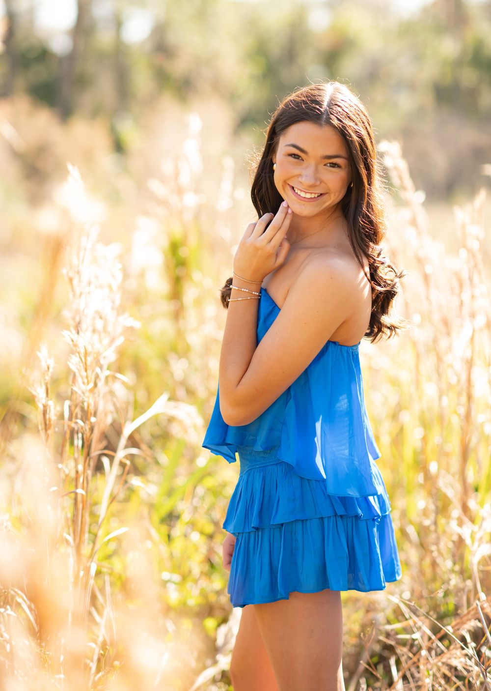 Smiling young woman in blue ruffled dress standing among golden tall grasses in a sunlit field.
