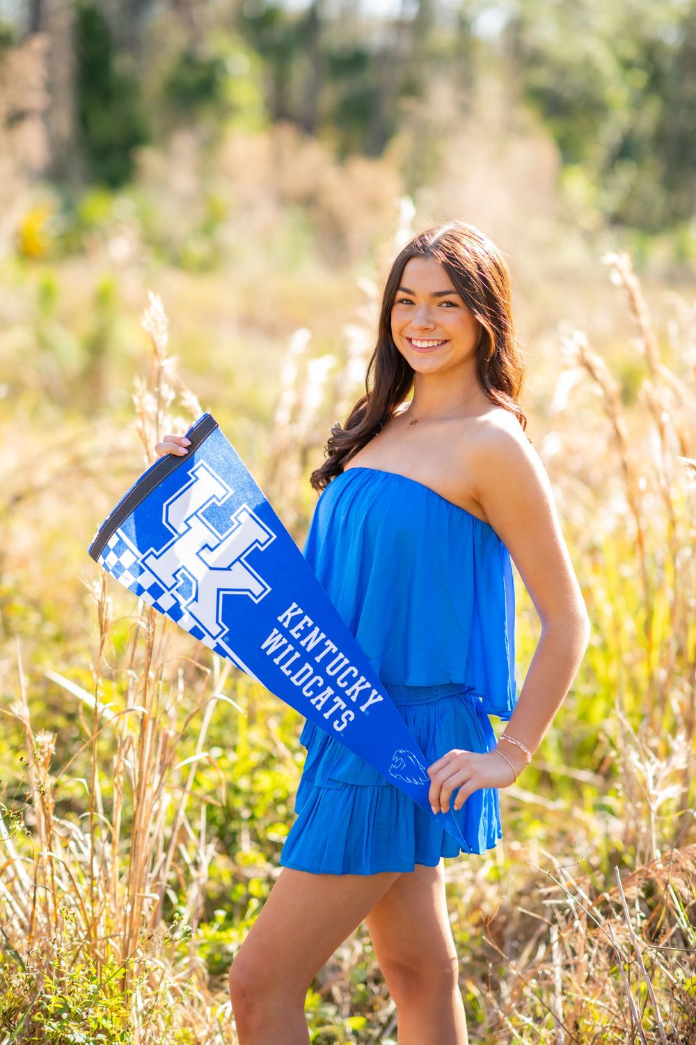 Smiling young woman in royal blue dress holding a Kentucky Wildcats pennant in a golden field.