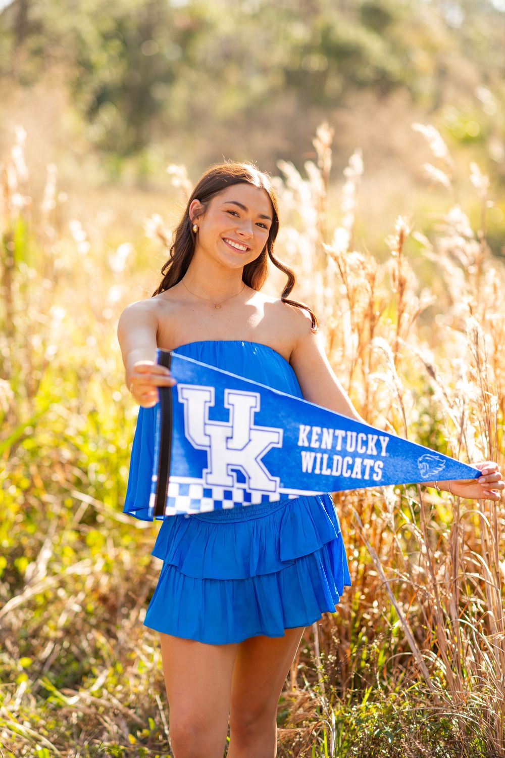 Smiling young woman in blue dress holding a Kentucky Wildcats pennant flag in a sunlit field.