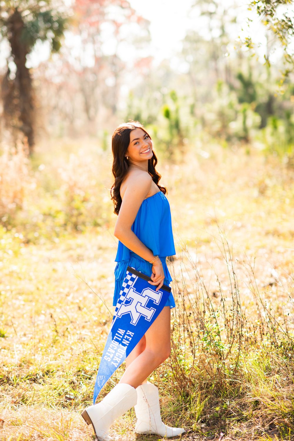 Smiling brunette woman in blue University of Kentucky outfit and white boots posing outdoors in a sunlit field.