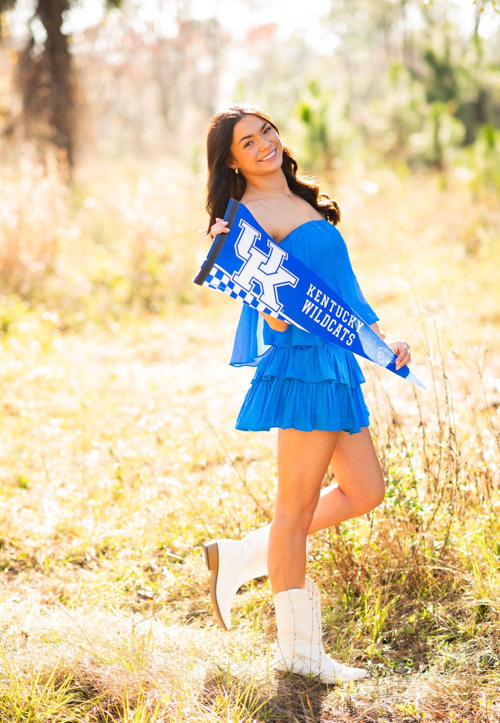 Smiling brunette woman in blue ruffle dress holding Kentucky Wildcats pennant flag outdoors in autumn field.