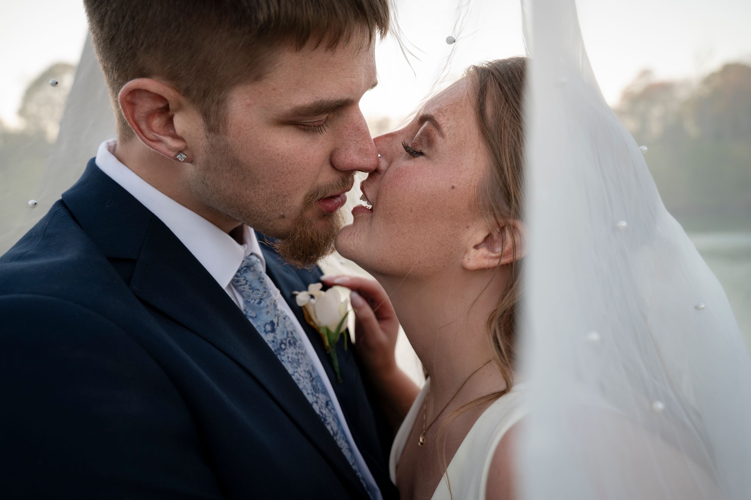 A bride and groom share a romantic kiss under a white veil on their wedding day outdoors.