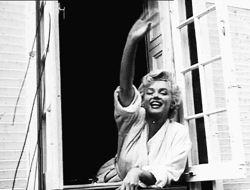 Black and white photo of a smiling woman with curly blonde hair waving from an open window while wearing a white robe.