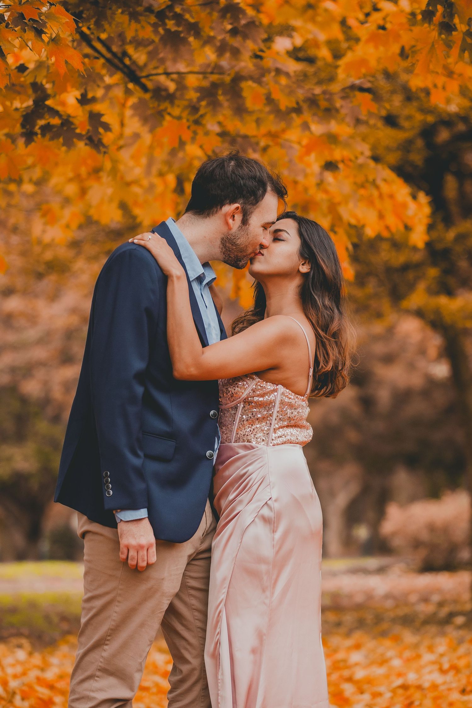 Couple in elegant outfits share a romantic kiss beneath vibrant orange autumn trees, with fallen leaves covering the ground in a park.