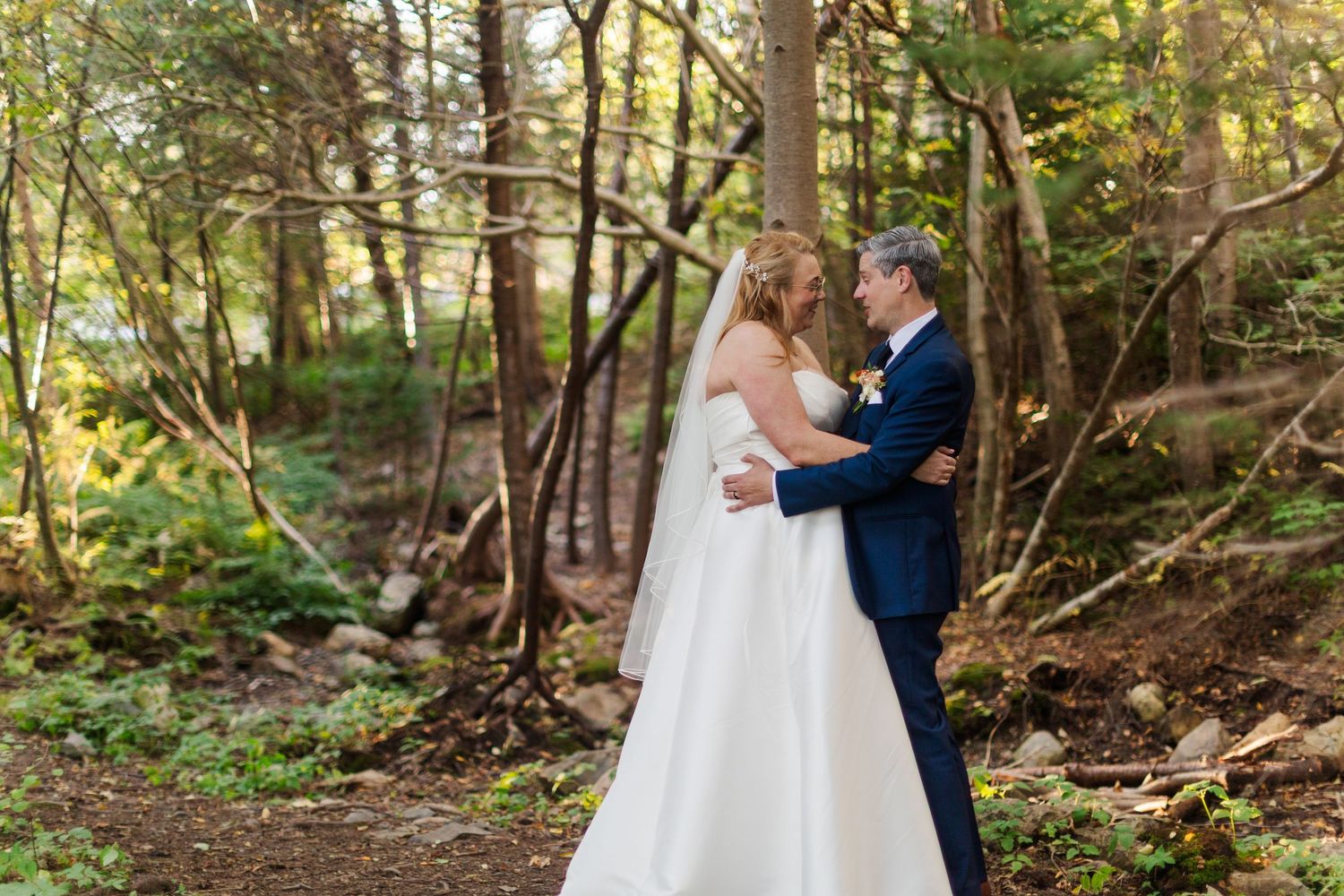 Bride in a white wedding gown and groom in a dark suit embrace in a sunlit forest clearing surrounded by tall trees.