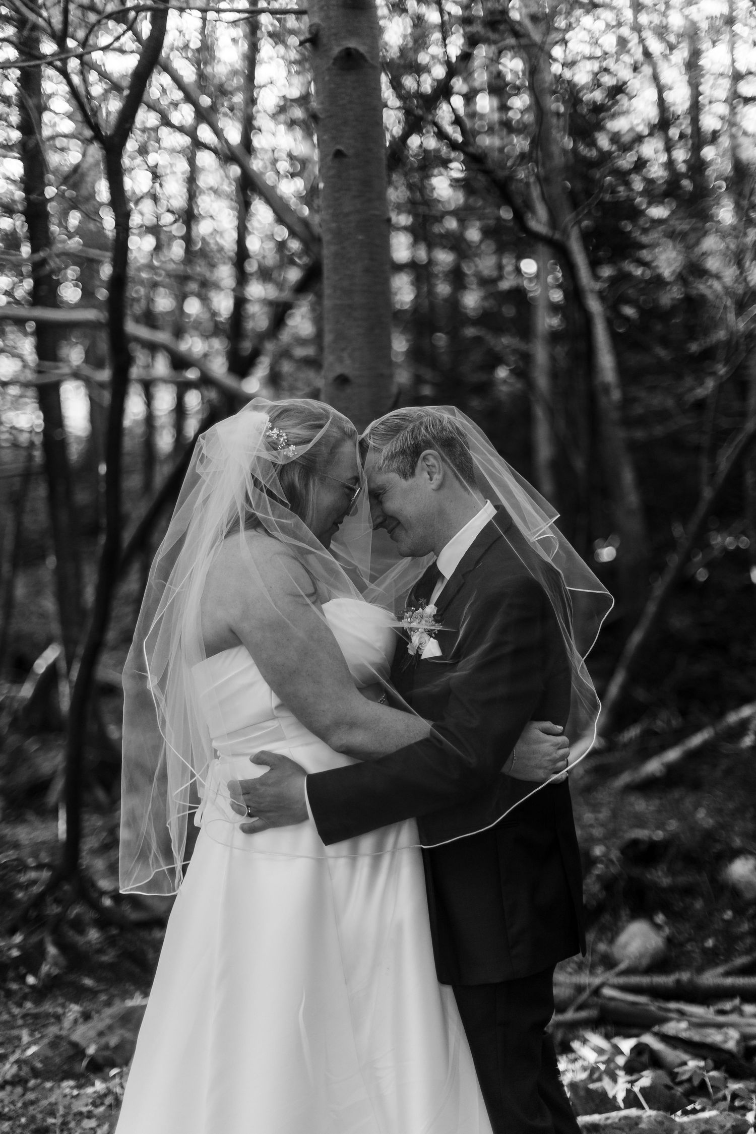 Black and white photo of a bride and groom embracing forehead to forehead under a veil in a quiet woodland setting.