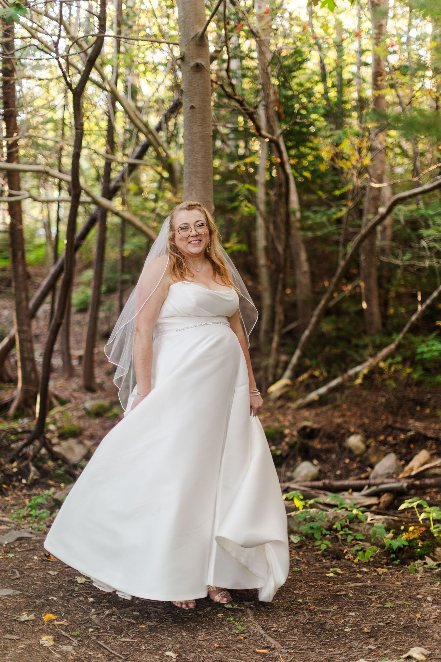 Smiling bride in a strapless white wedding gown and sheer veil stands on a leafy forest path surrounded by tall trees.