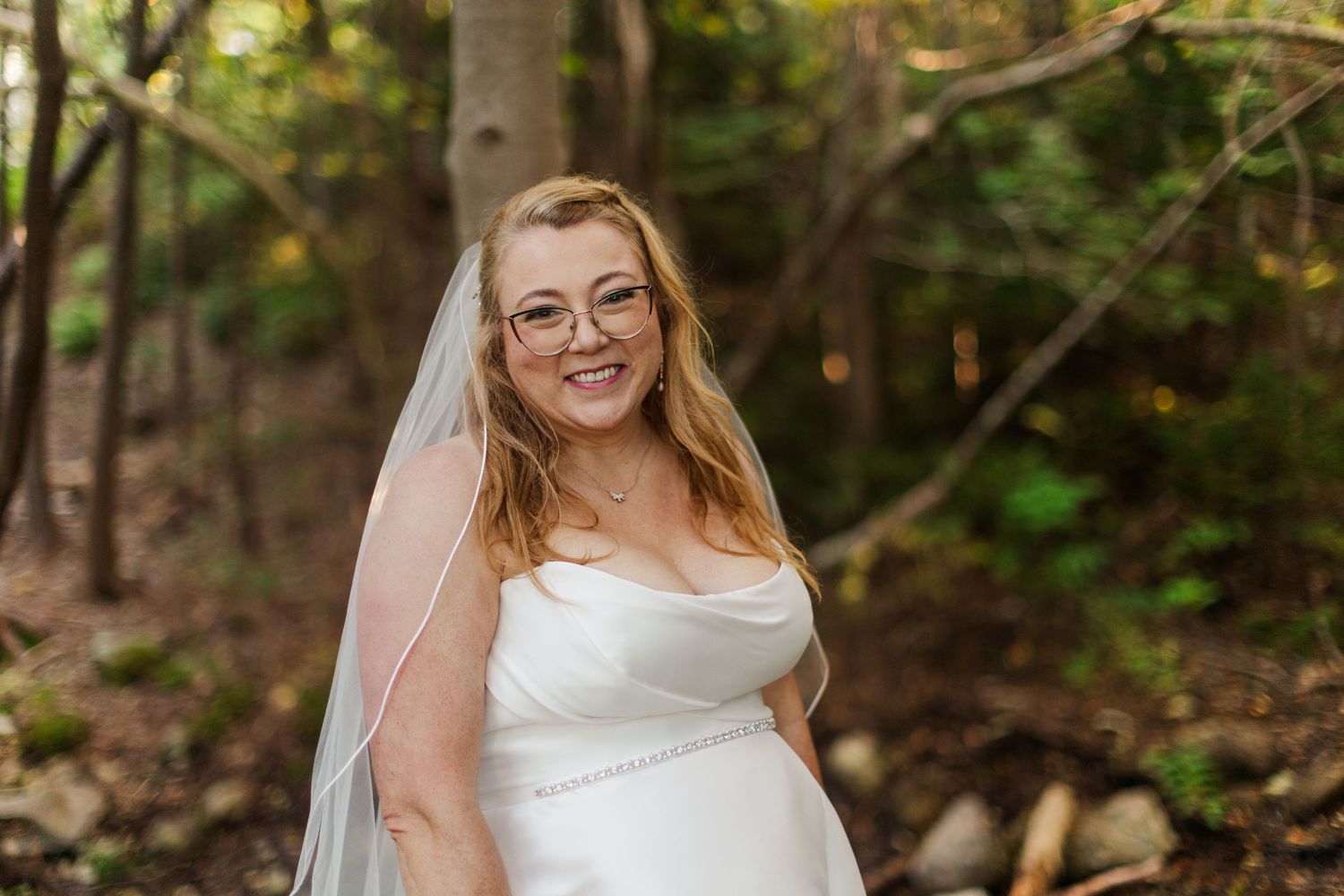 Smiling bride in glasses wearing a white satin wedding dress and sheer veil, posing outdoors in a sunlit wooded setting.