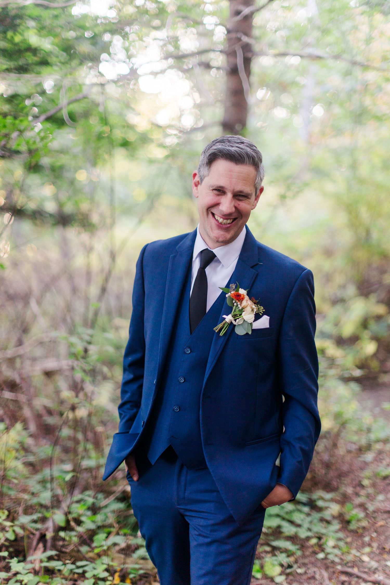 Smiling groom in a navy blue three-piece suit with a floral boutonniere and dark tie stands outdoors among trees in a wooded setting.