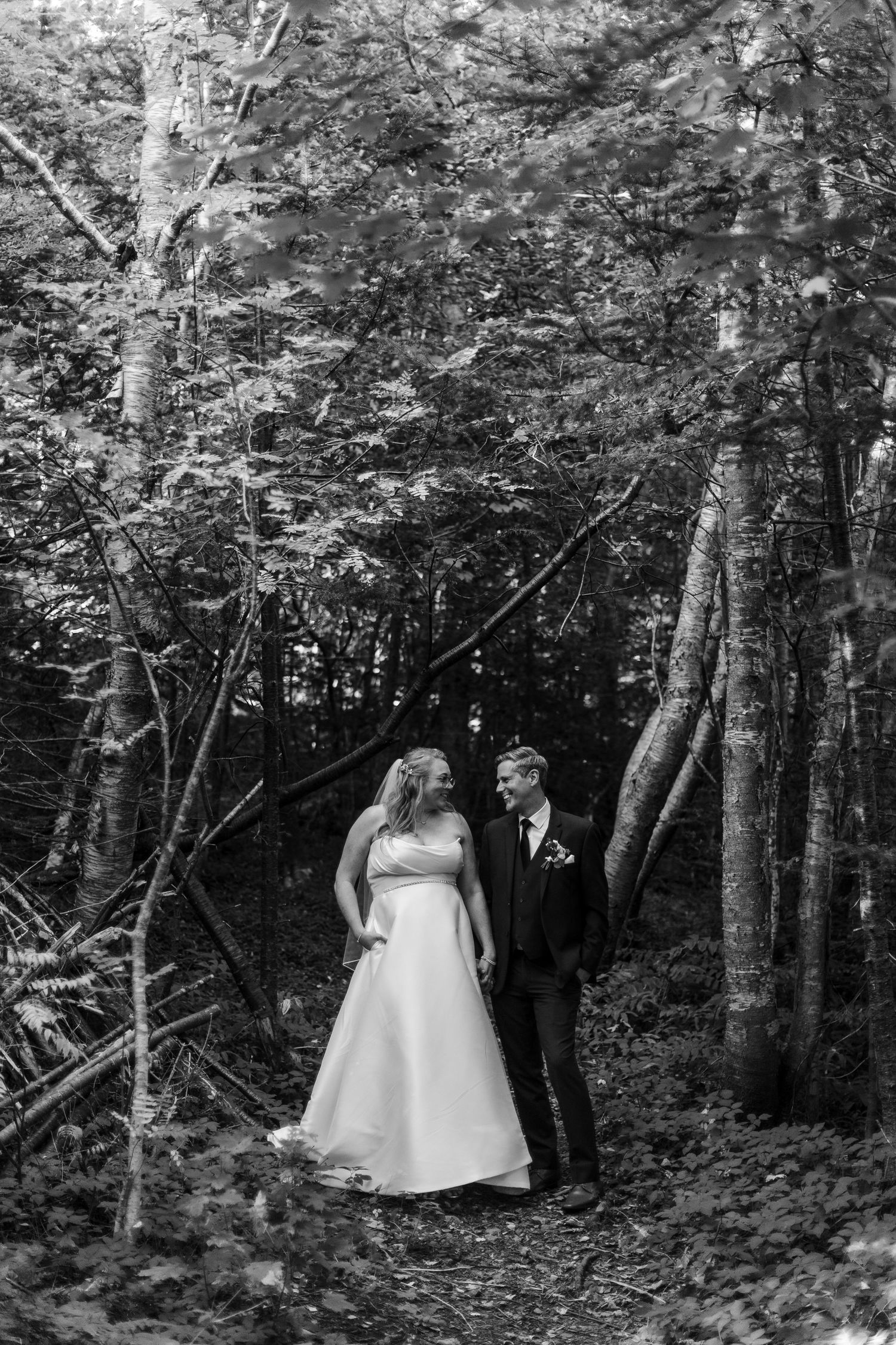 Black and white photo of a smiling bride and groom sharing a happy moment on a wooded trail among tall trees.