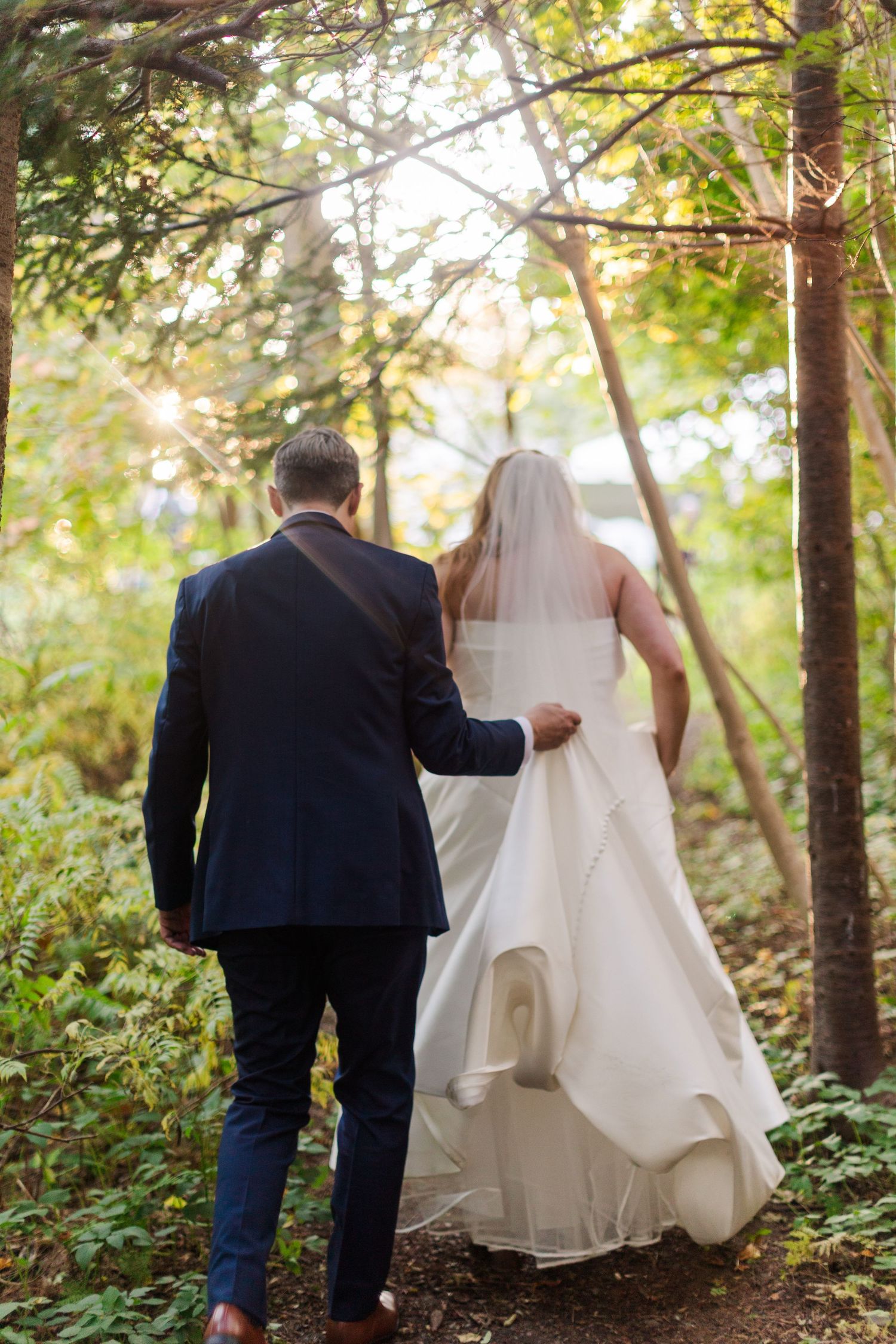Groom in a navy suit leads his bride in a flowing white wedding gown along a sunlit forest path.