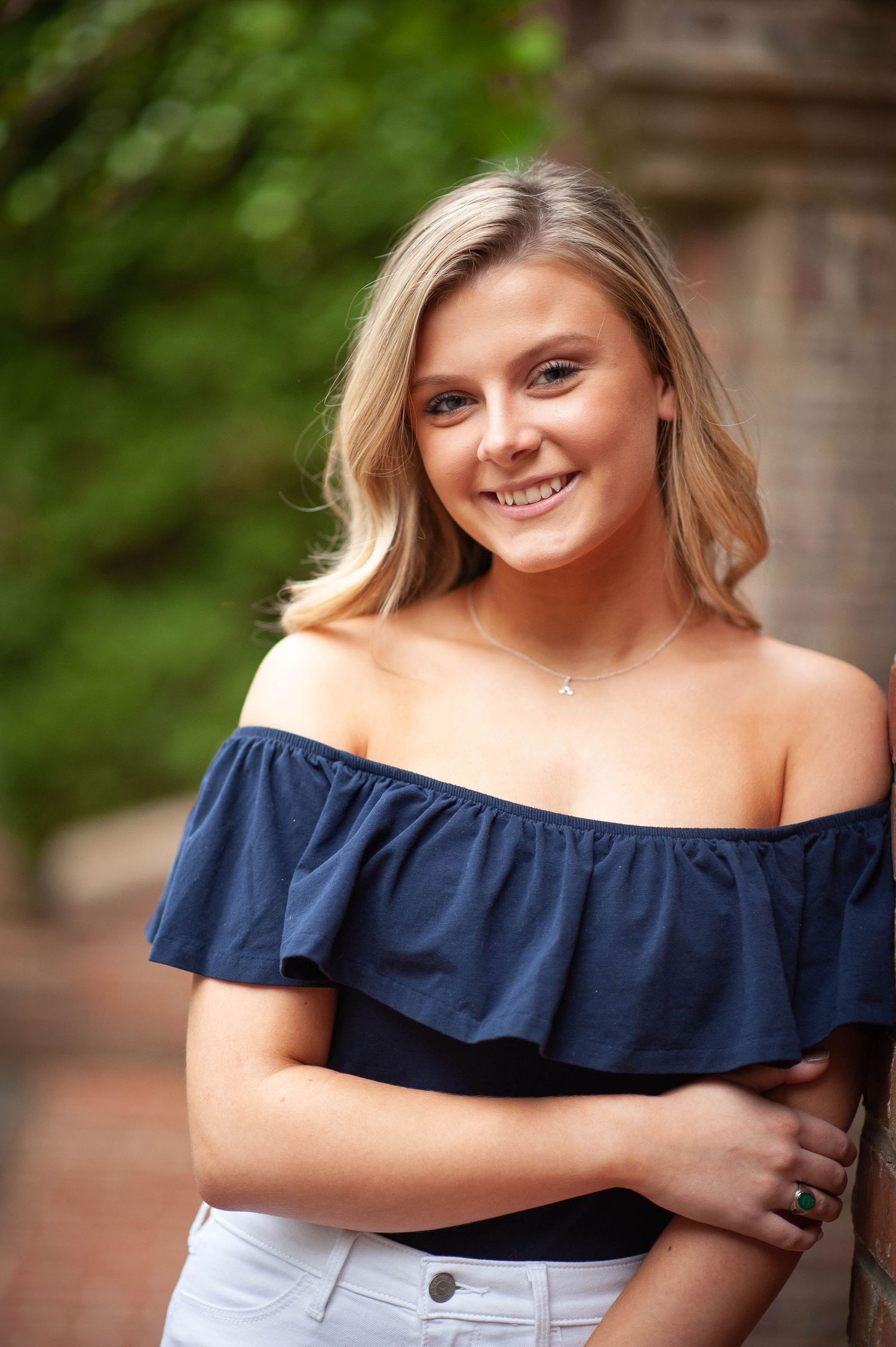 Smiling young blonde woman in navy off-shoulder top and white jeans posing outdoors near brick wall.