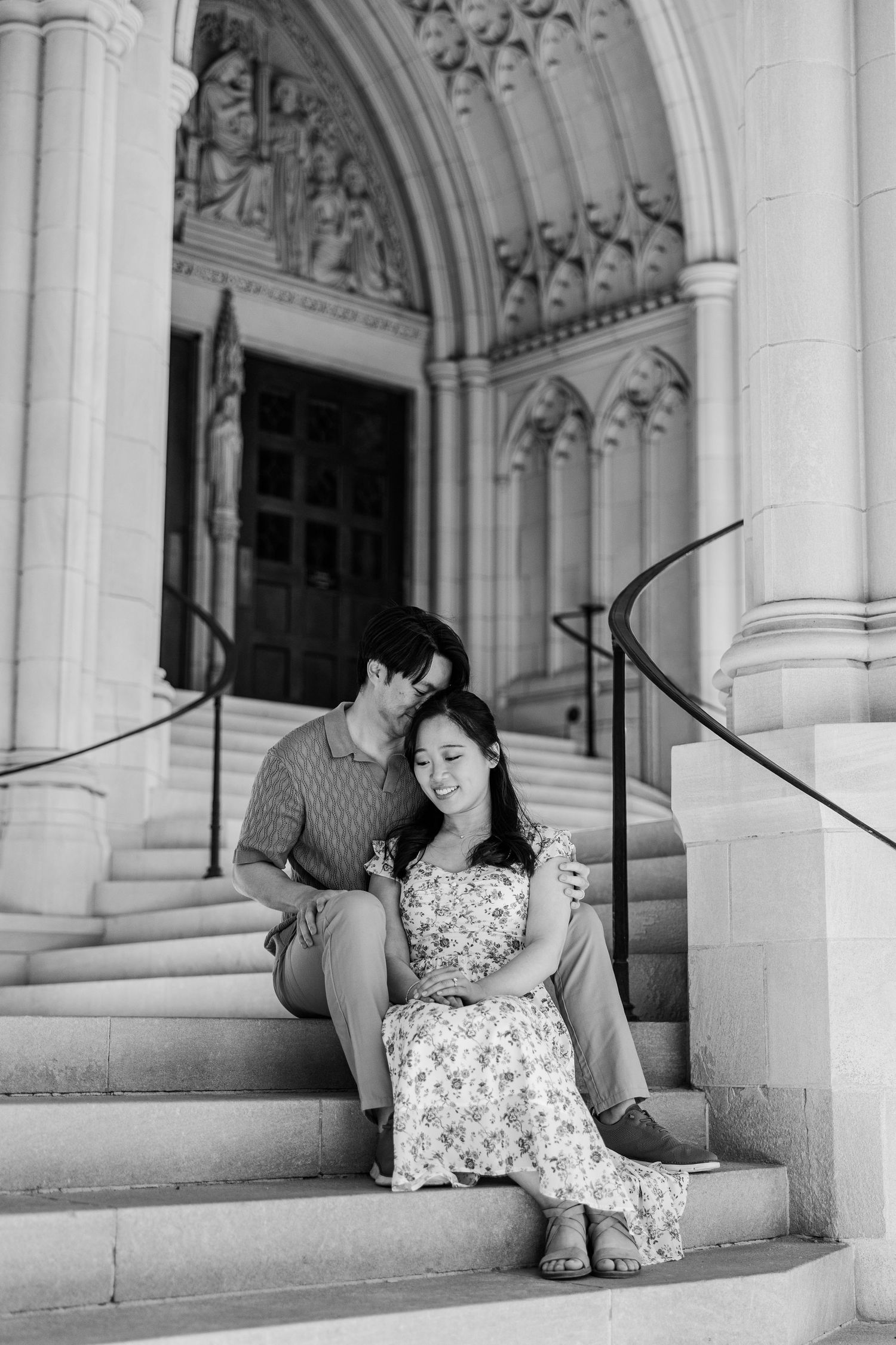 Black and white photo of a couple sitting on church steps, woman in floral dress, man leaning in close, grand arched entrance behind them.