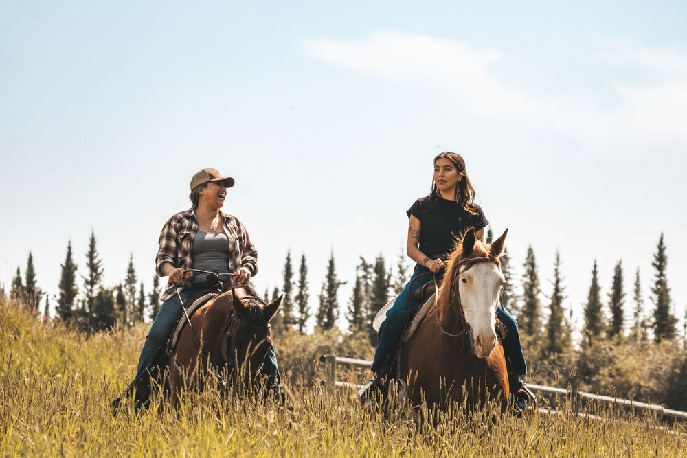 Two women riding horses through a sunny golden field with evergreen trees in the background.