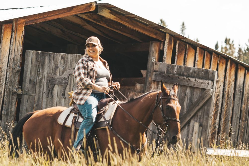 Woman in plaid shirt and cap riding a chestnut horse in front of a rustic wooden barn.