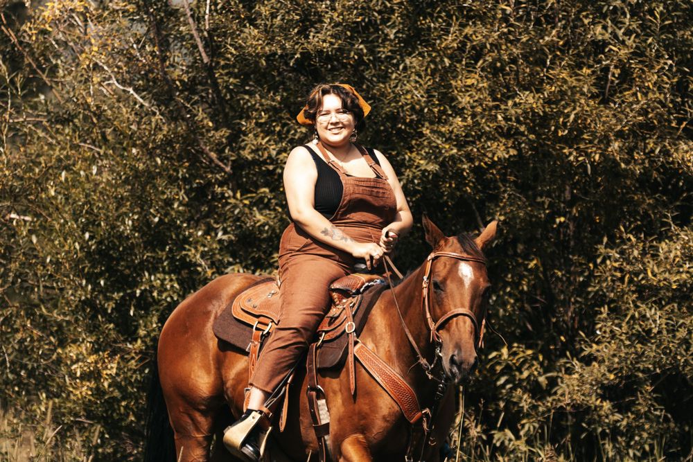 Woman in brown outfit smiling while riding a chestnut horse outdoors.