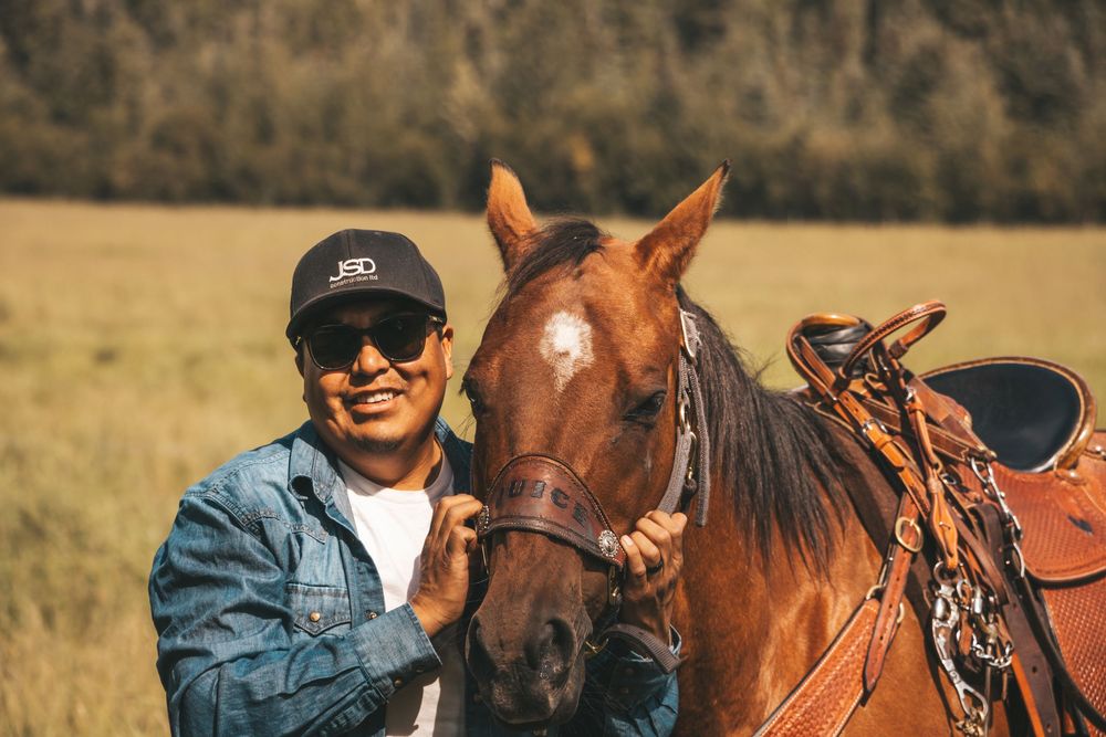 Man in denim jacket and cap smiling beside a saddled brown horse in a scenic meadow.