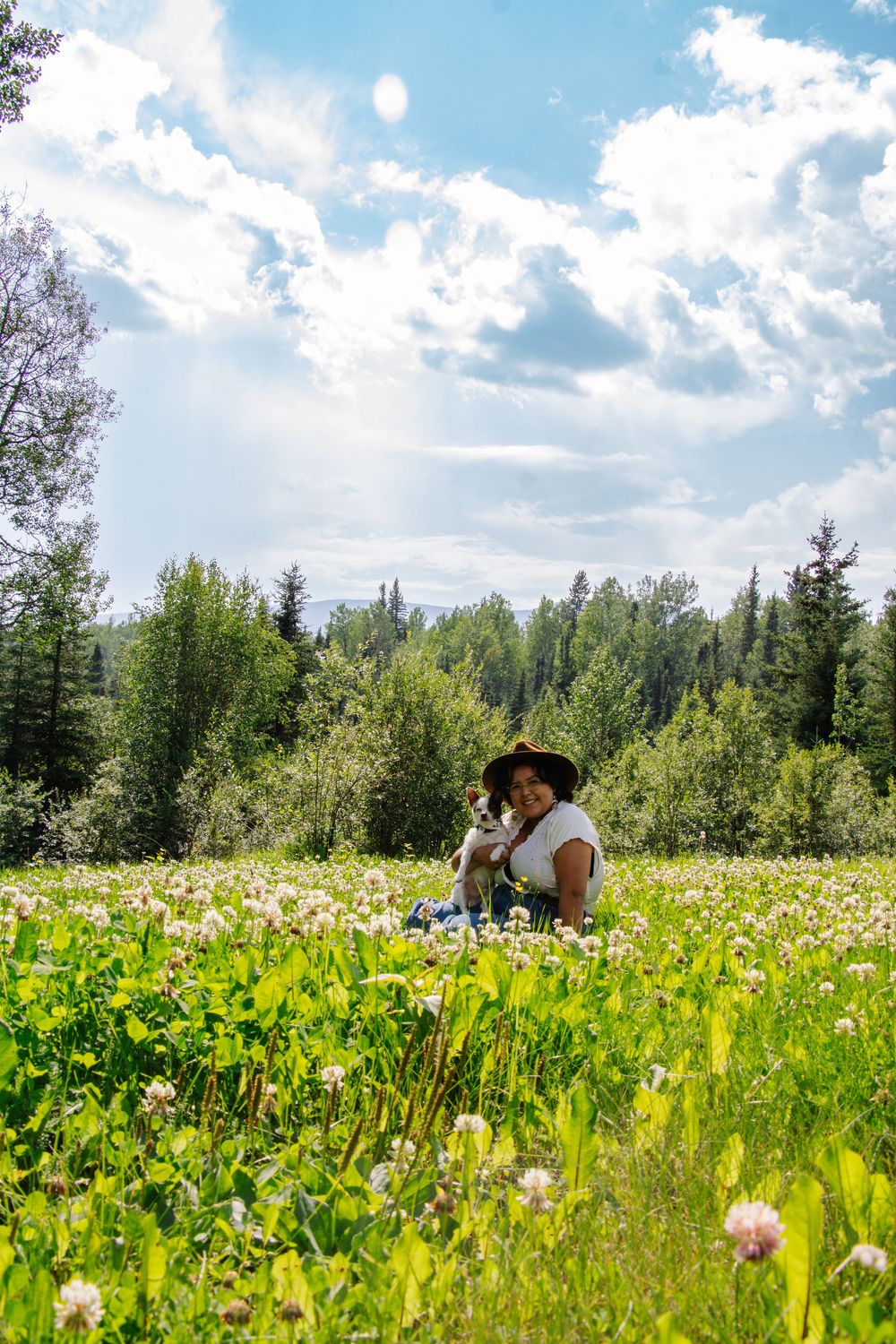 Bride in white dress and hat holds a dog in a sunny wildflower meadow.