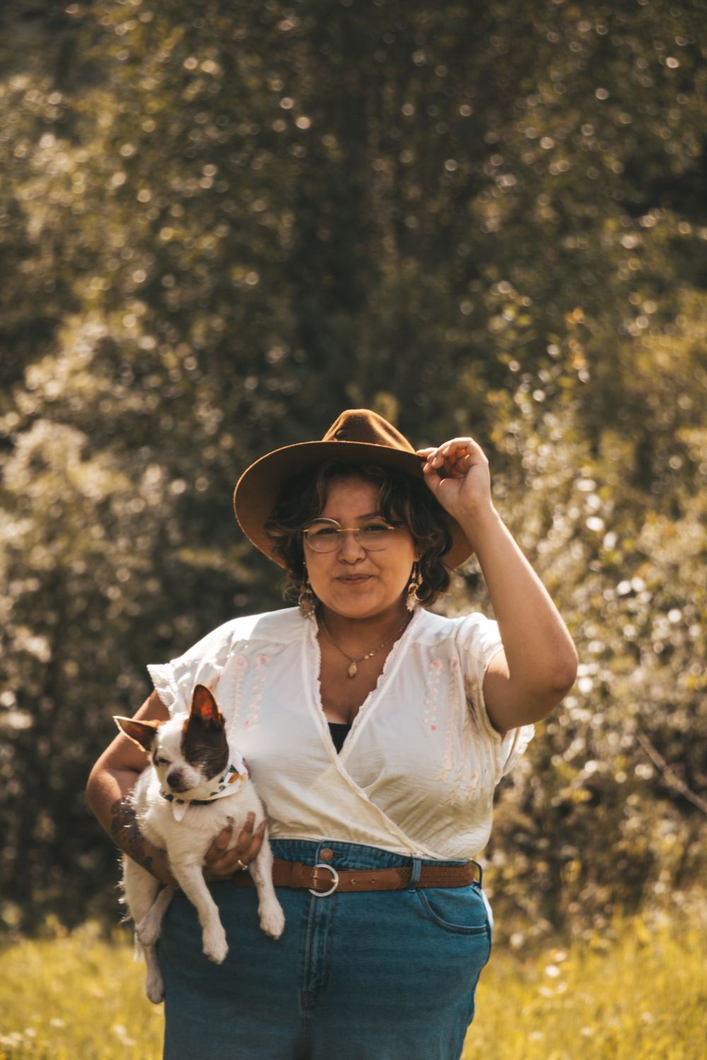 Woman in brown hat and white blouse holds small dog outdoors in sunny green field.