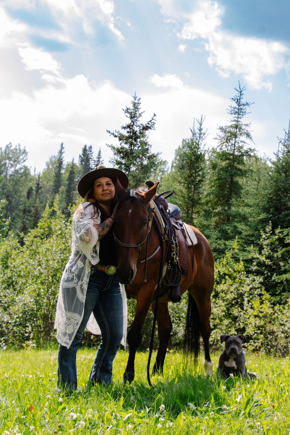 Woman in cowboy hat with brown horse and black dog in lush green meadow with pine trees and cloudy sky.