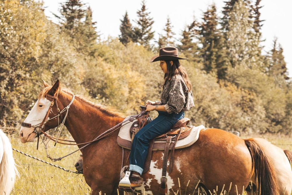 A woman in a cowboy hat rides a chestnut horse through a green meadow with trees in the background.
