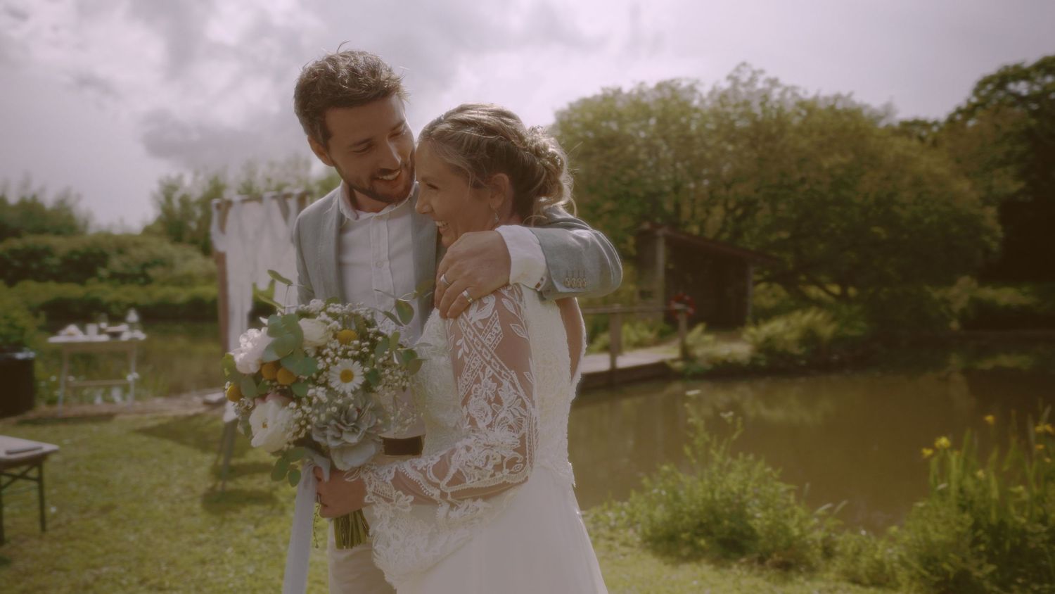 A joyful couple embraces outdoors on their wedding day, bride holding a bouquet near a serene garden pond.