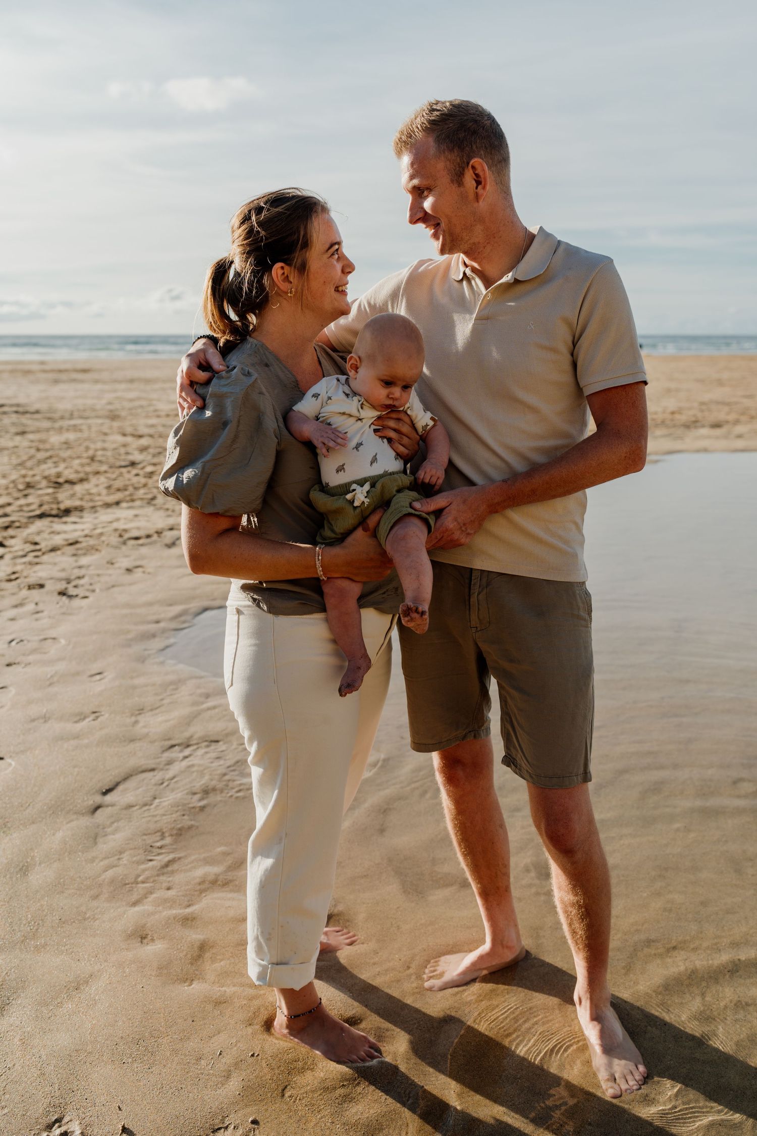 Happy family with baby enjoying a sunny beach day, parents smiling at each other while holding their infant on sandy shore.