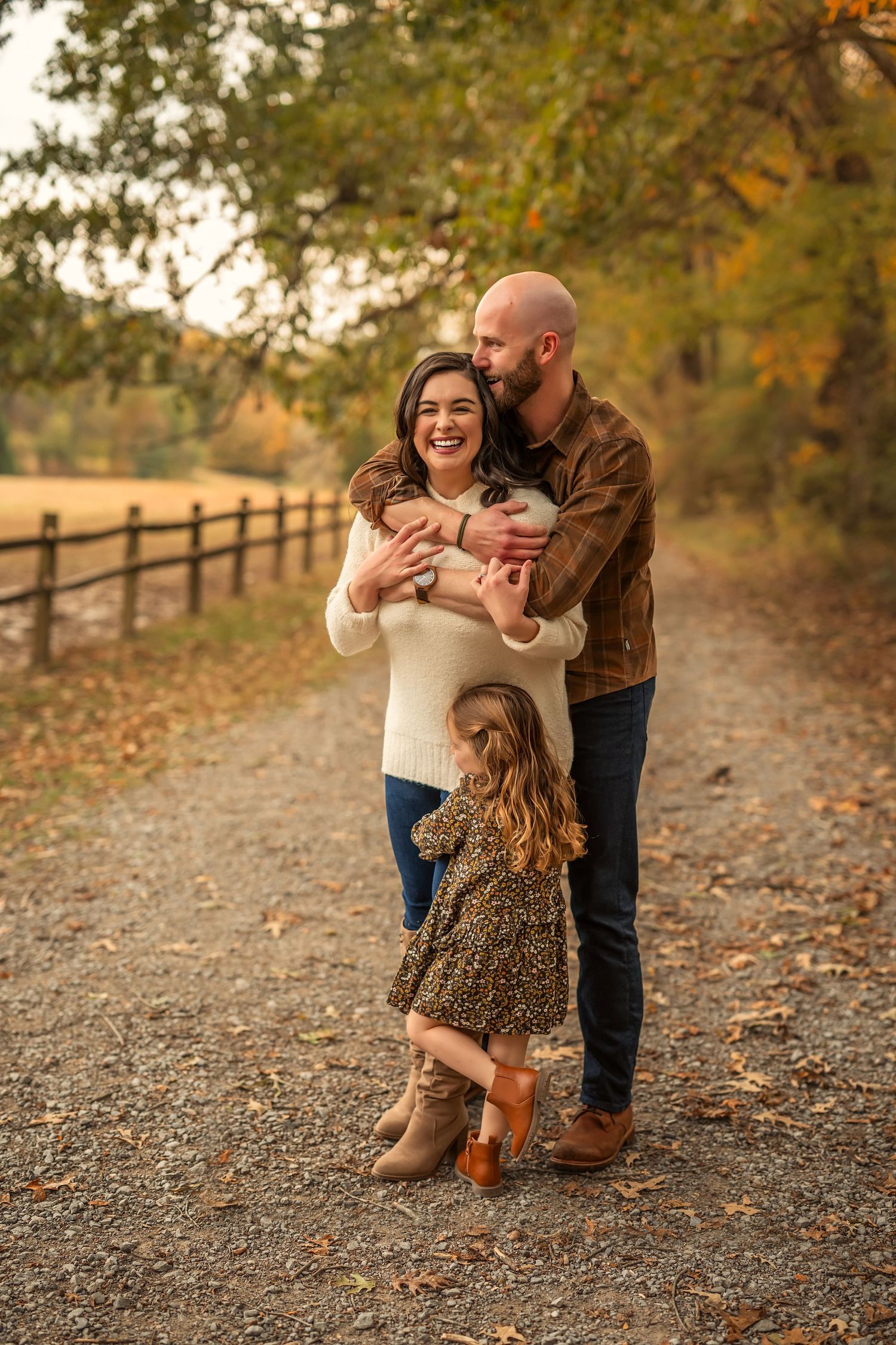 Family of three poses on a rustic path surrounded by autumn foliage, parents embracing while young daughter stands in front.