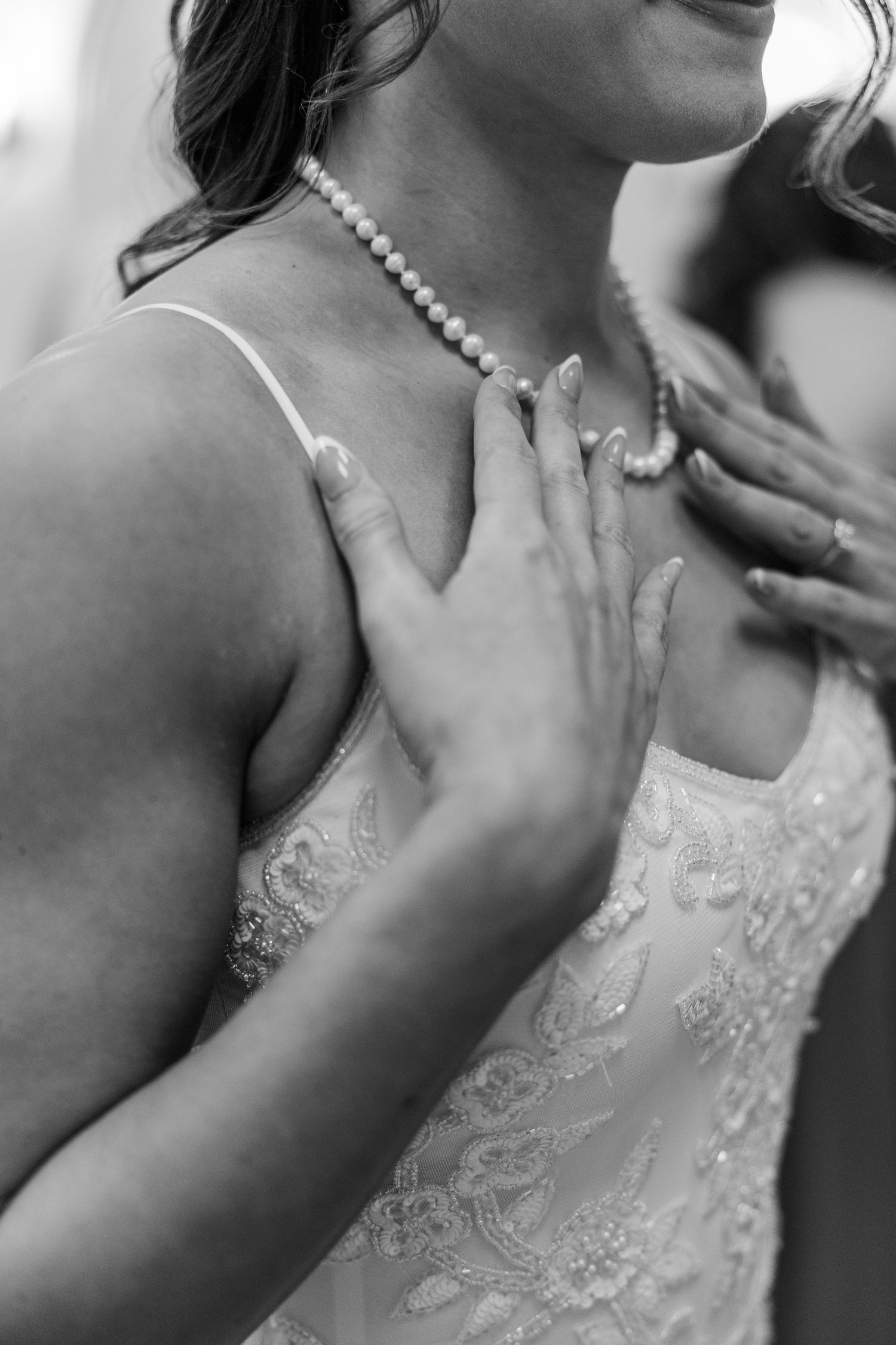 Black and white photo of a bride in lace dress having pearl necklace fastened by another person.