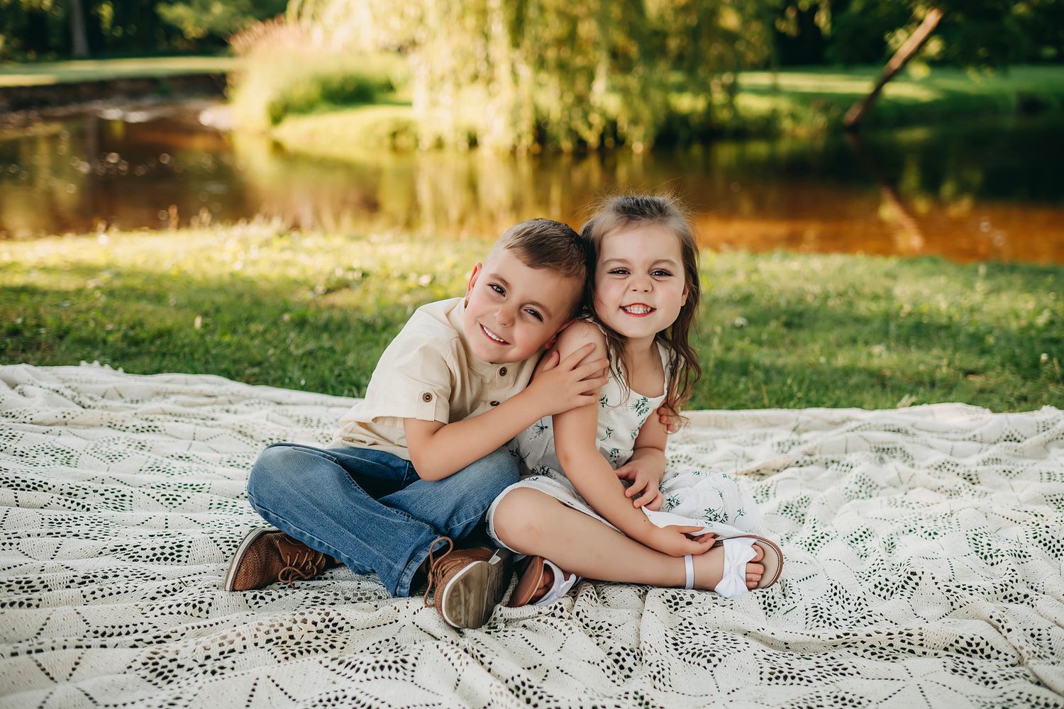 Two young siblings hugging and smiling on a blanket outdoors near a pond on a sunny day.
