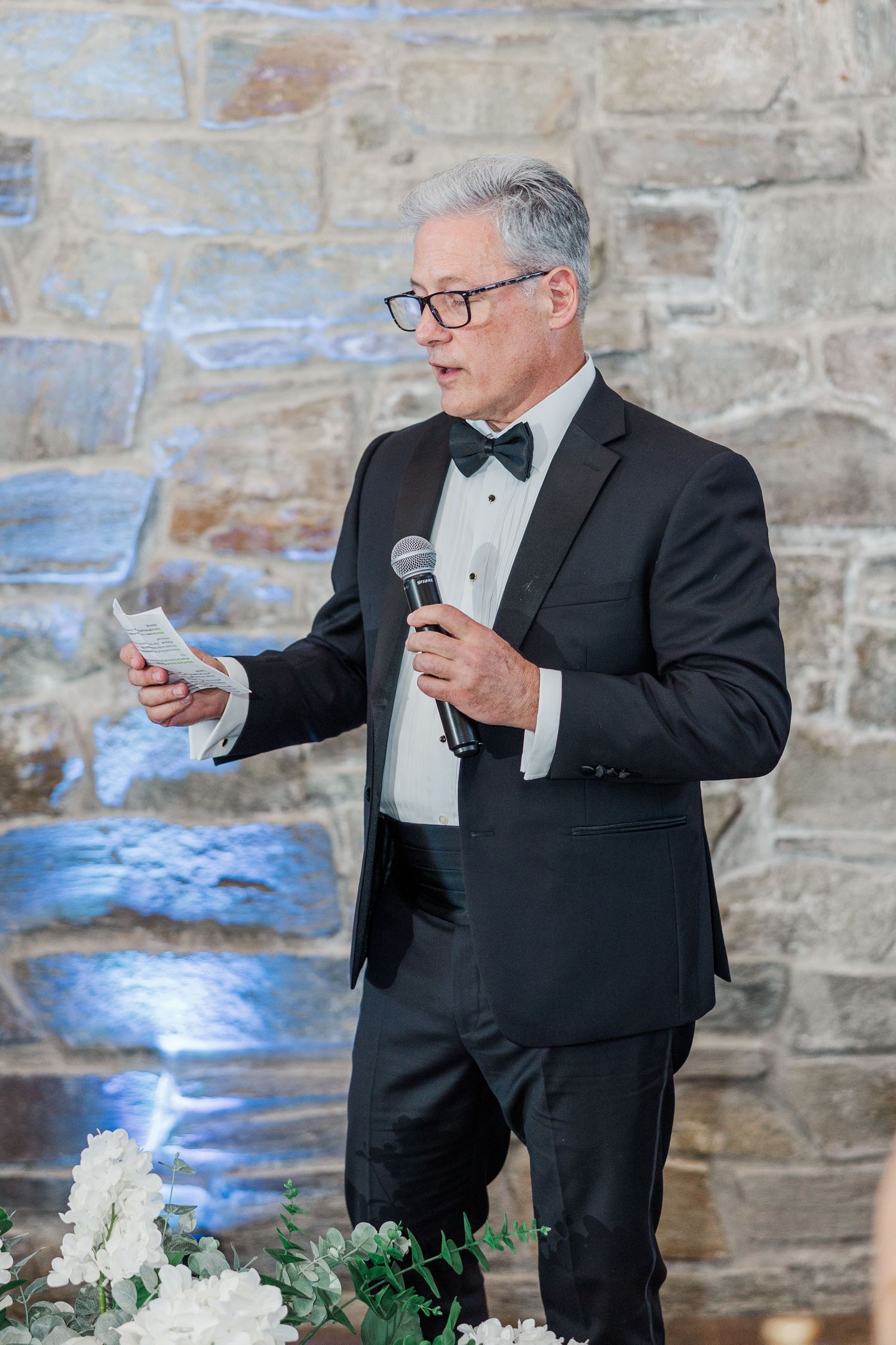 Elegant man in tuxedo delivering wedding speech at reception, holding microphone and notes near stone wall.