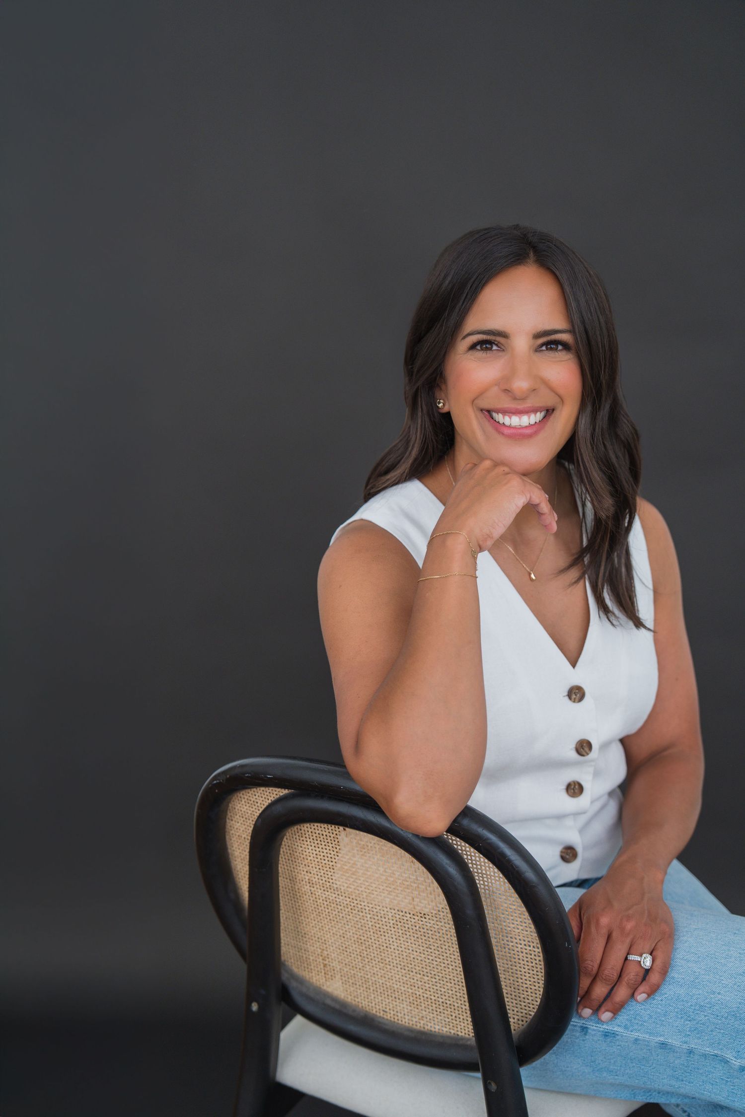 Smiling brunette woman in white sleeveless top and jeans sitting on a chair against a dark gray background.