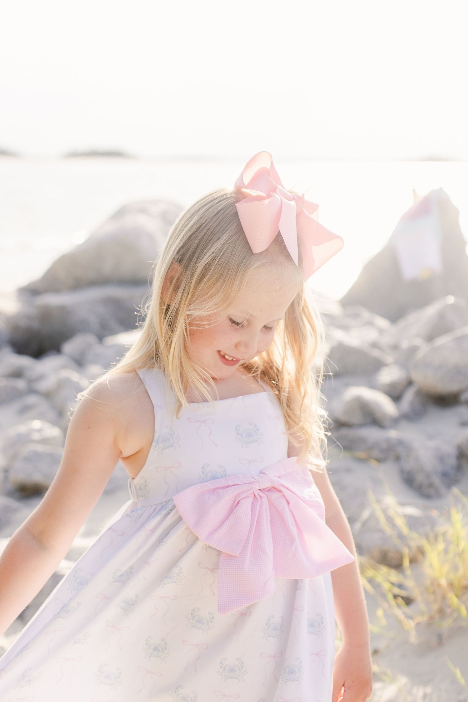 Young girl at Fripp Island Beach, showing off her white dress with pink bow at waist and hair bow, smiling at rocky beach in soft light.
