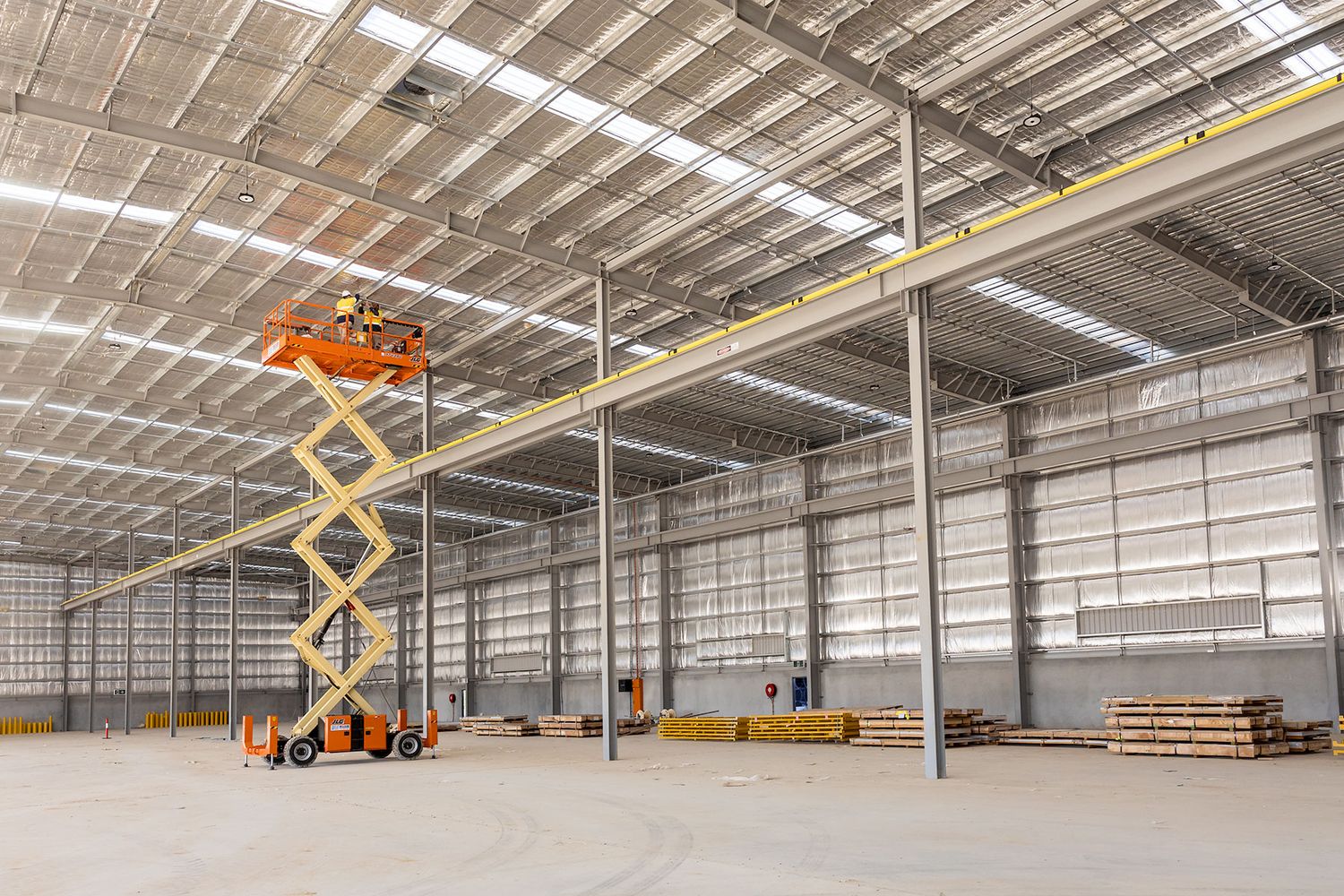 Orange scissor lift in a large industrial warehouse under construction with steel frame ceiling.