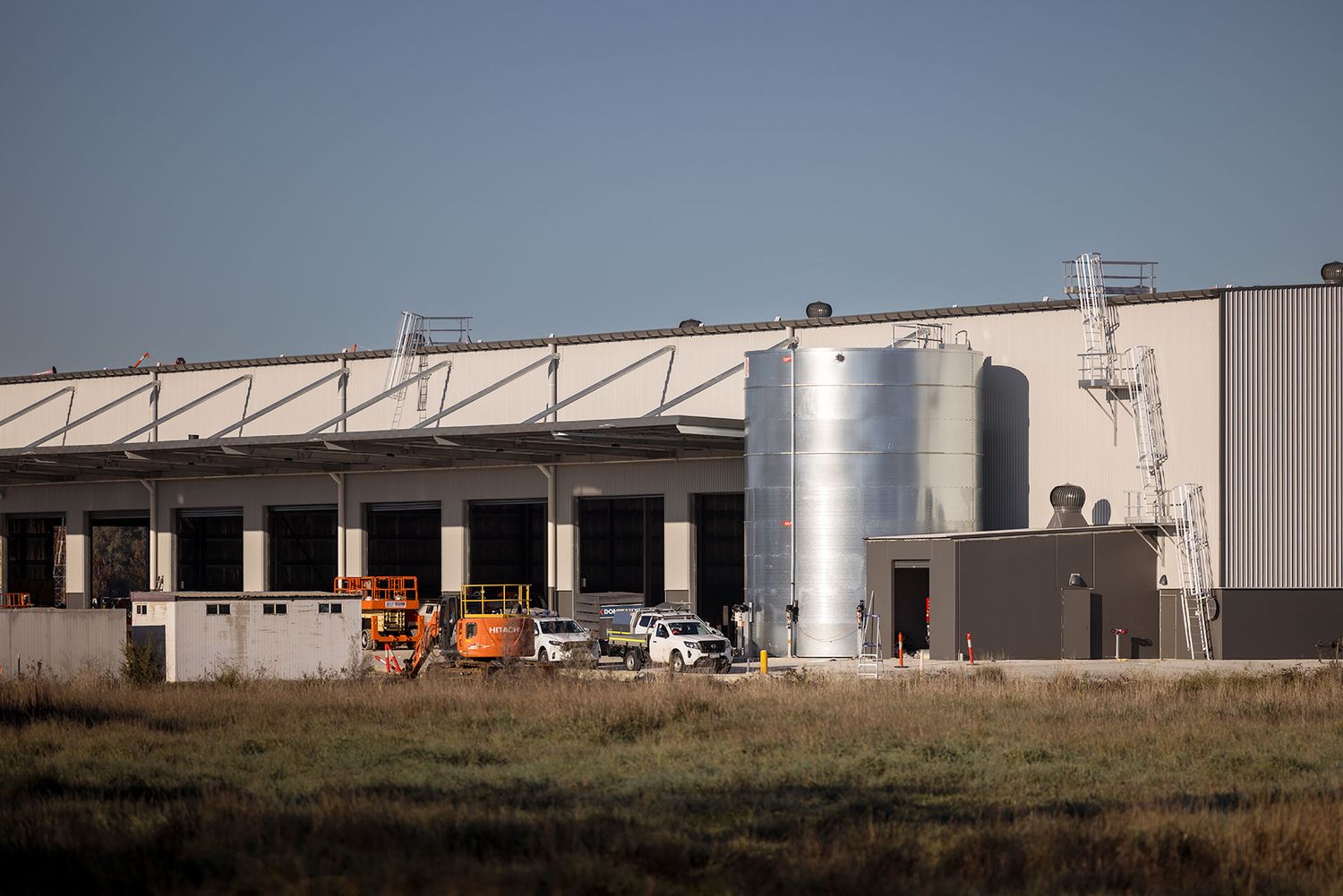 Industrial warehouse facility with silver storage silos, loading docks, and construction vehicles on flat terrain under clear sky.