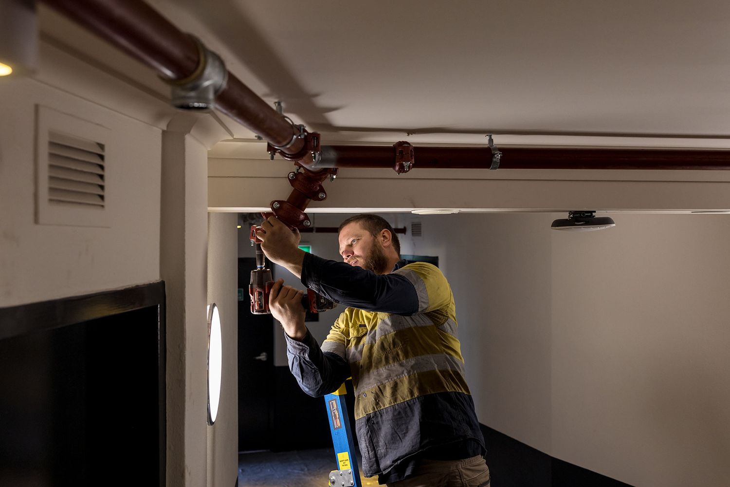 A worker in a safety vest installs water pipes for a fire sprinkler systemon a ceiling in a dimly lit corridor.