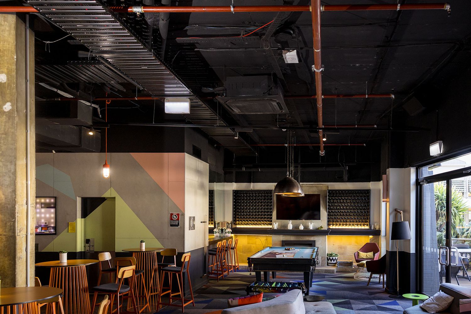 Modern industrial bar lounge with exposed black ceiling, pool table, pendant lights, and colorful geometric wall accents.