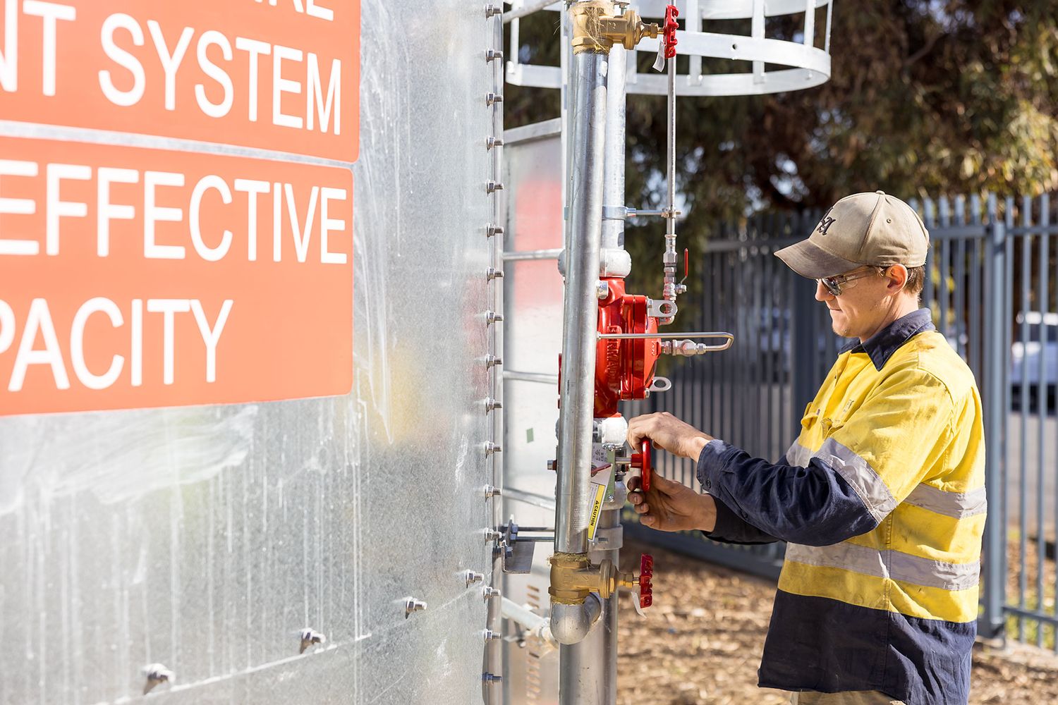 Worker in hi-vis clothing inspecting industrial tank system valves at an outdoor facility set up for fire protection.
