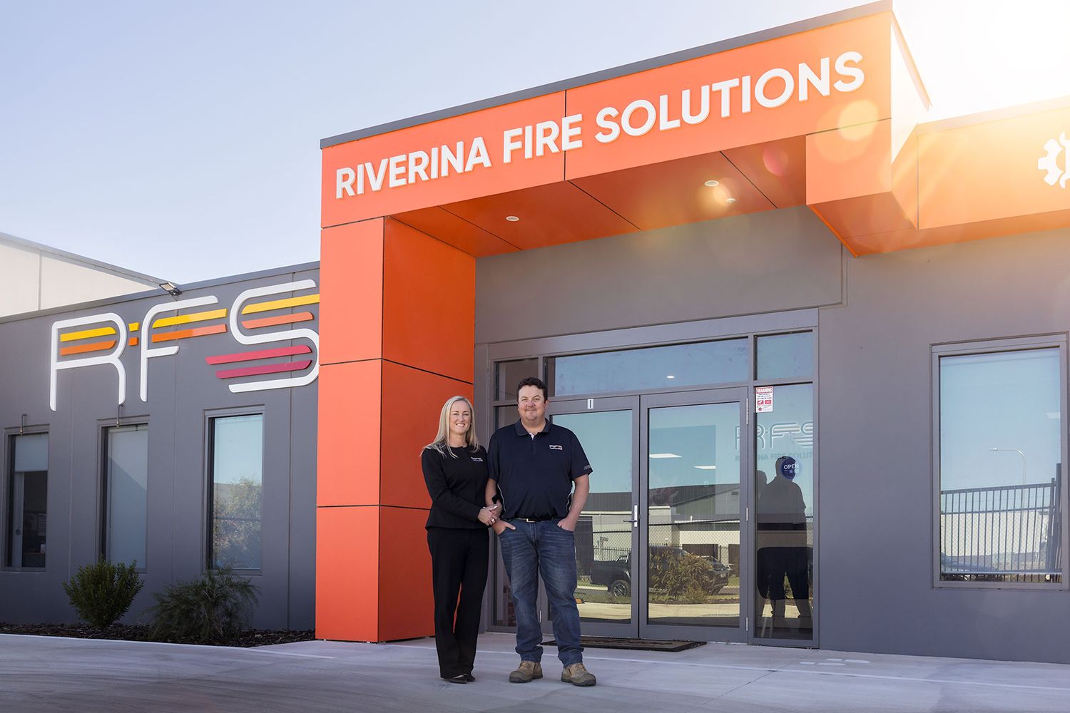 Two staff members stand outside Riverina Fire Solutions modern building with bold orange RFS signage at sunset.