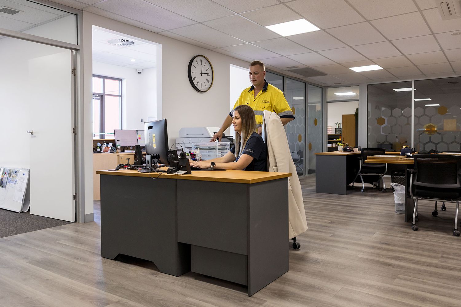 Two office workers at a reception desk, one in a yellow shirt standing, one seated at a computer in a modern open-plan office.