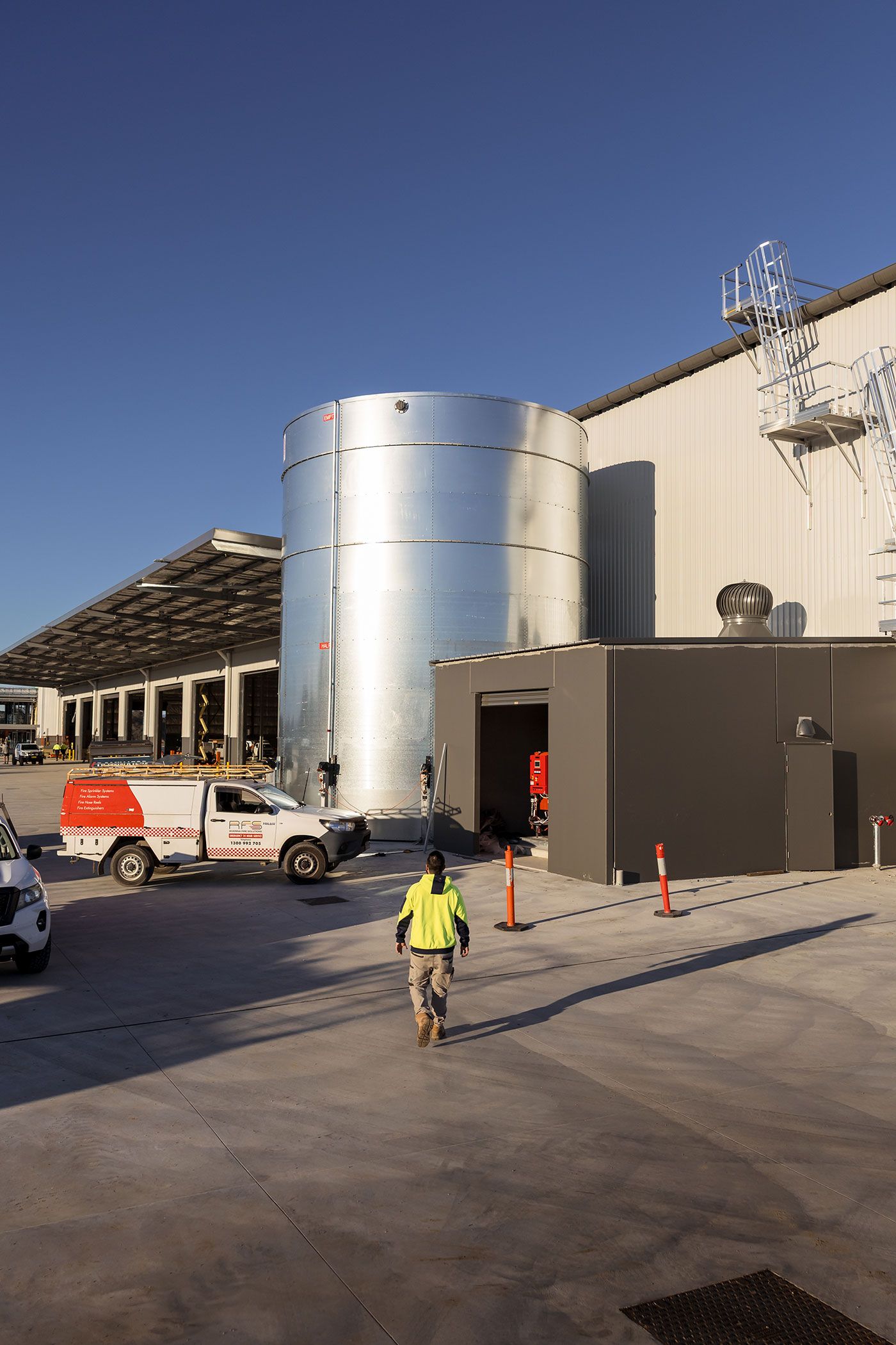 Worker in hi-vis vest walks toward large silver industrial tanks at a construction site under a clear blue sky.