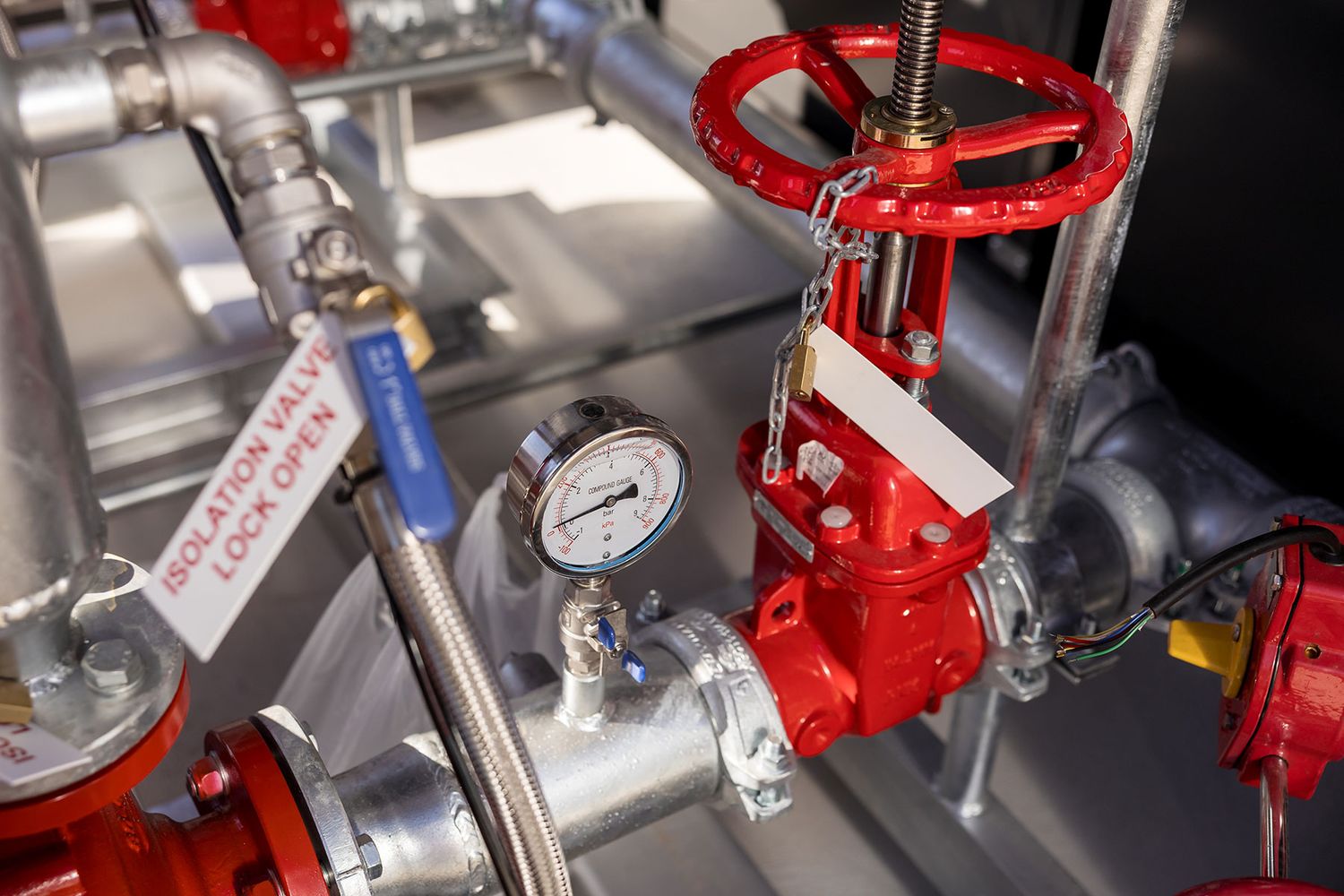 Red industrial gate valve with pressure gauge on fire suppression system pipes in mechanical room.