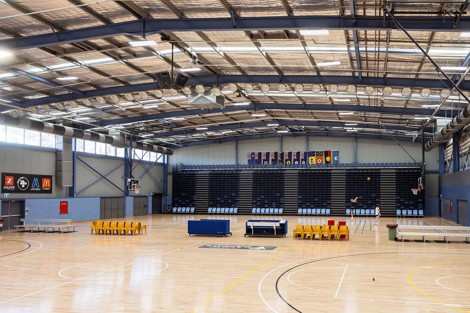 Large indoor sports gymnasium with hardwood basketball court, bleacher seating, and high ceiling with natural lighting.