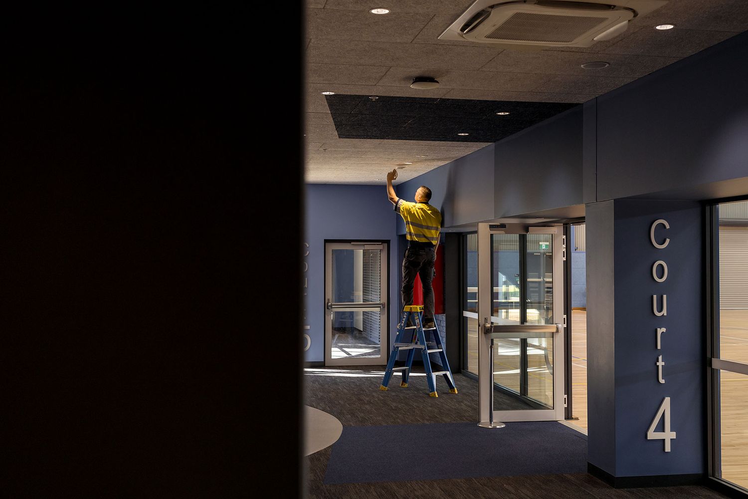 Worker on ladder performing maintenance near Court 4 entrance in a dimly lit indoor sports facility.