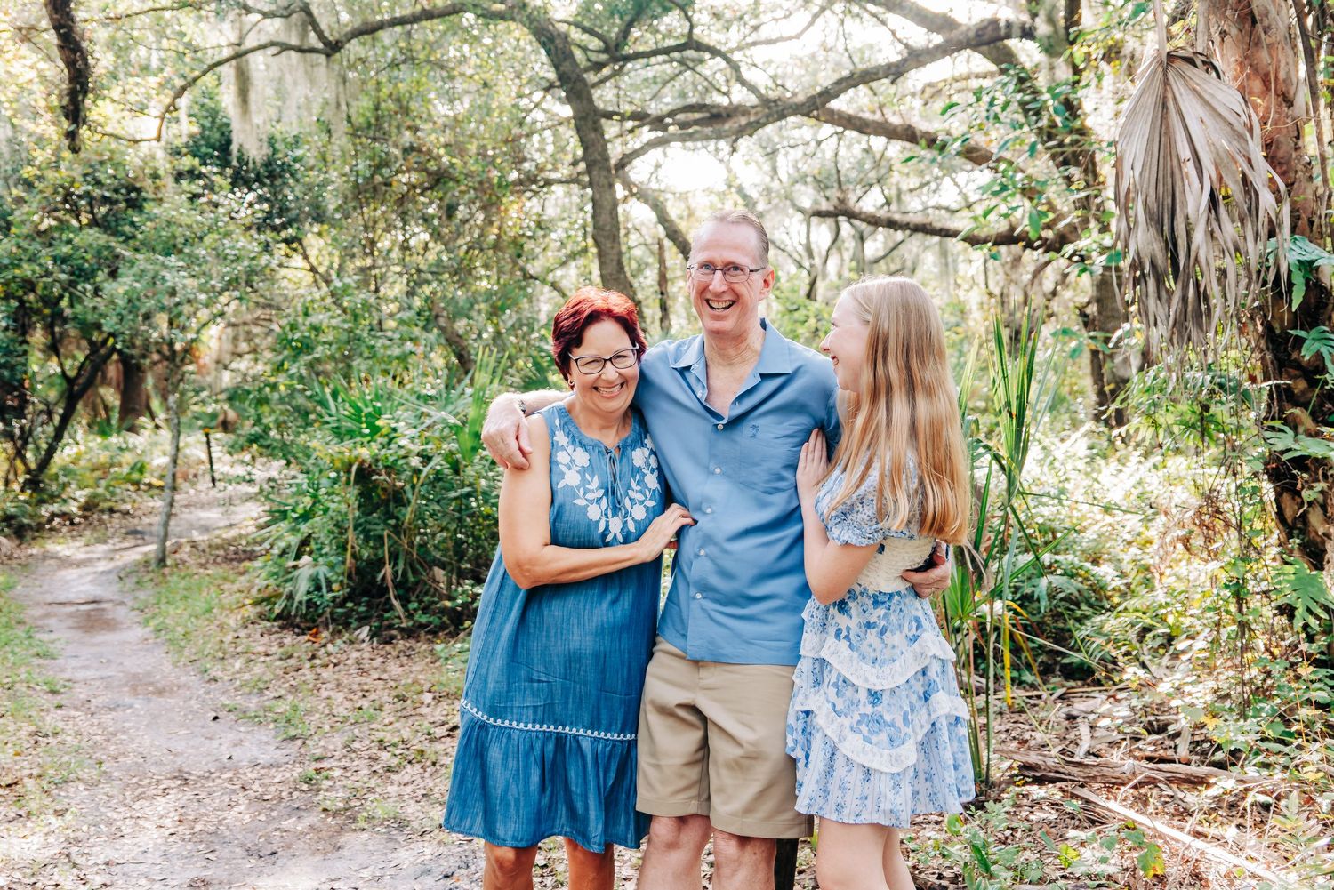 Family of three smiling together on a scenic wooded path, dressed in coordinating blue outfits at Delray Oaks Nature trail Boca Raton FL.