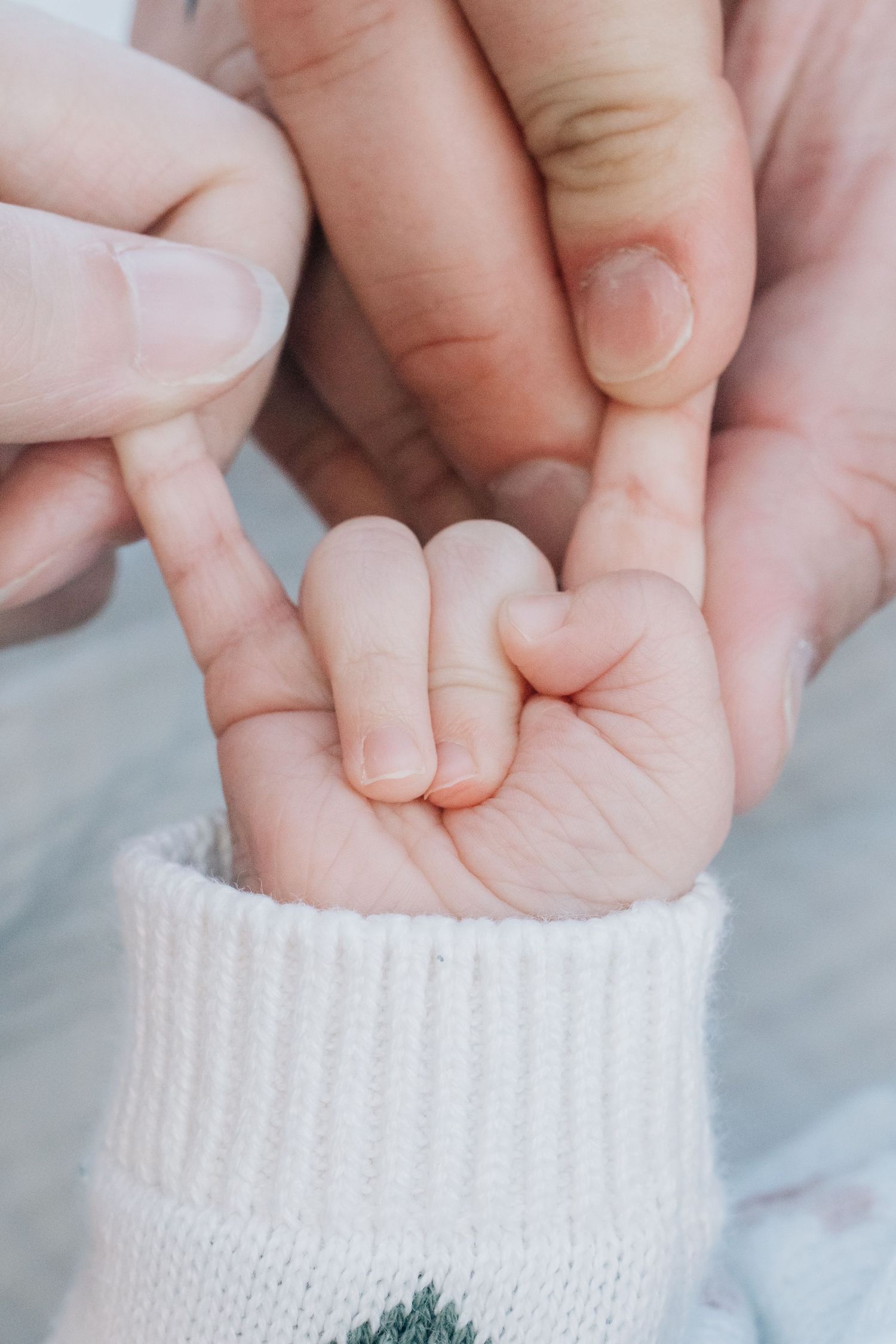 Close-up of a newborn baby's tiny hand grasping an adult's finger, wearing a white knit sleeve.